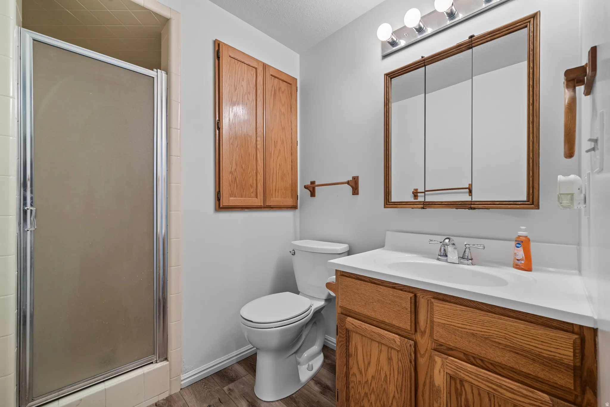 Bathroom featuring a vanity with a white countertop and wood-finish cabinetry, a framed medicine cabinet with integrated lighting, a toilet, a built-in wall cabinet, and a shower with a frosted glass enclosure