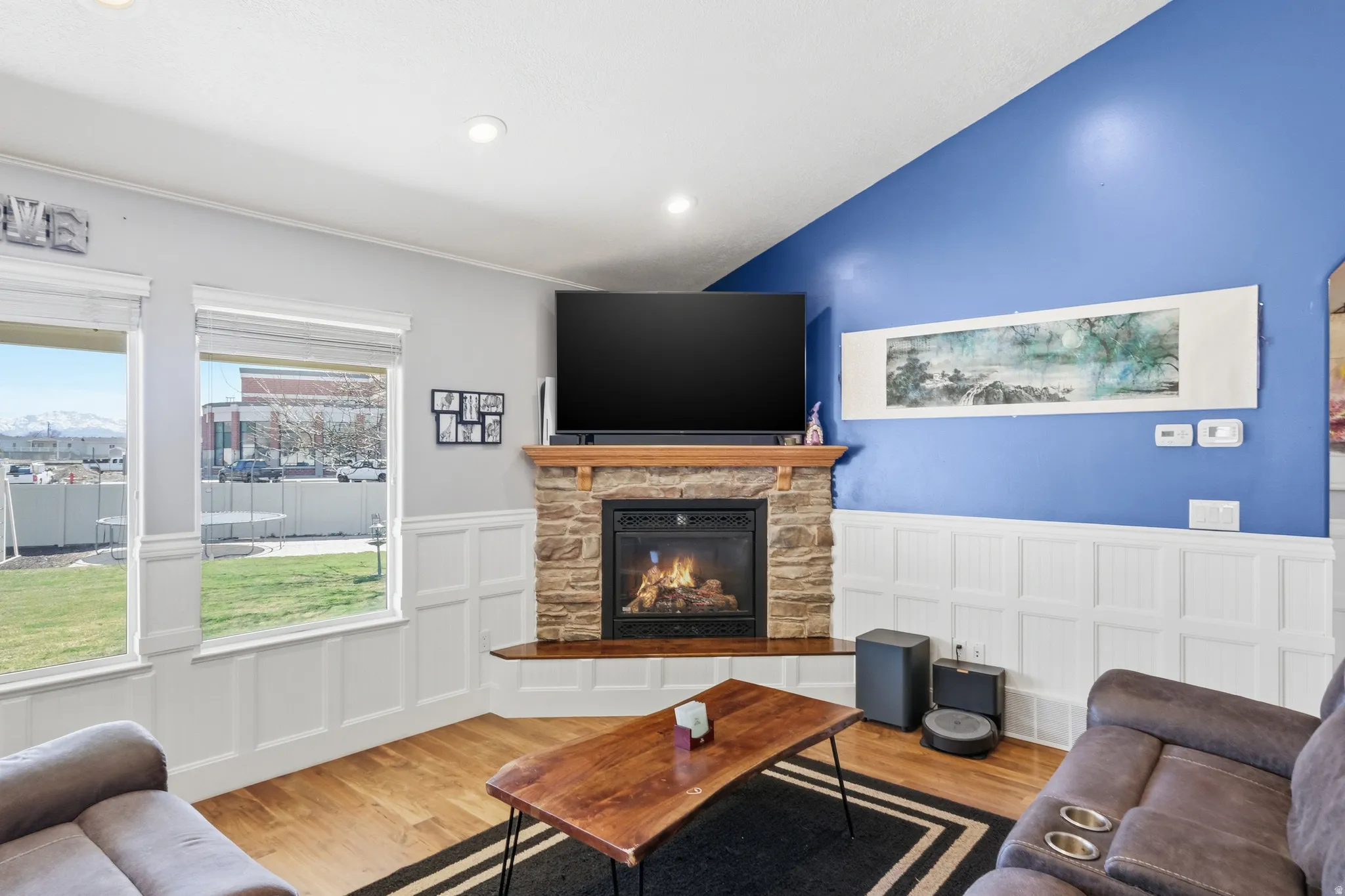 Stone-clad fireplace with a wood mantle and hearth, recessed lighting, wood-finish flooring, two large windows, and white wainscoting