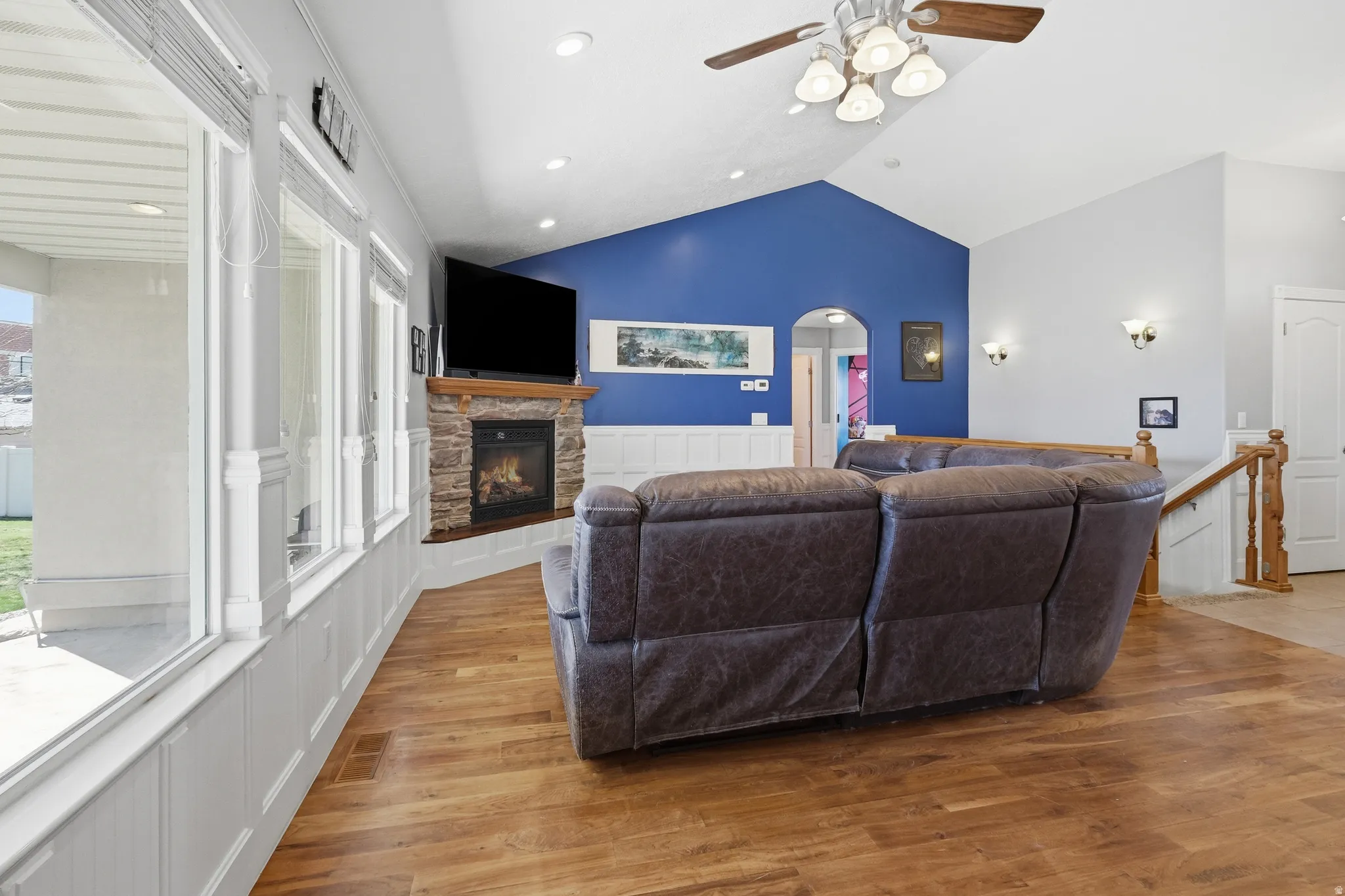 Vaulted ceiling living area featuring wood-finish flooring, a stone-surround fireplace with a wood mantle, and a wall of windows above wainscoting