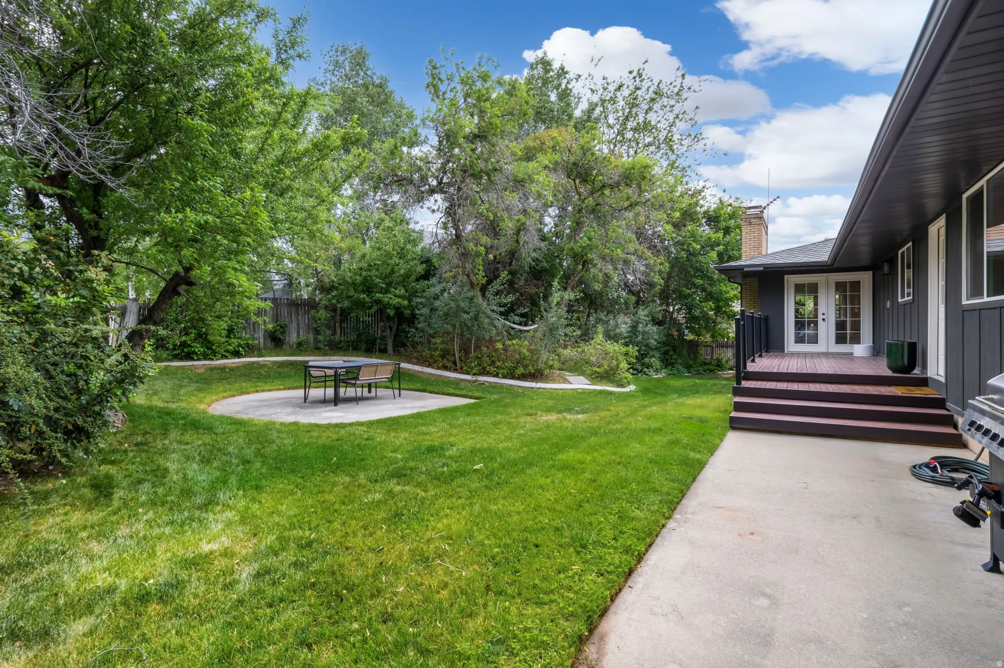 Expansive backyard featuring a manicured lawn, a circular concrete patio, mature trees, a multi-level wood deck, and French doors leading into the property