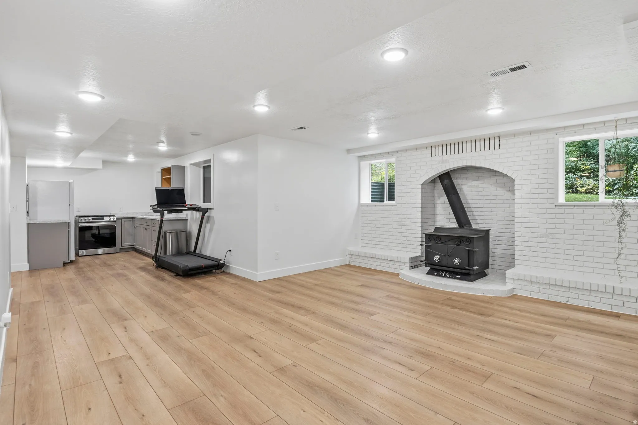 Spacious open-concept living area featuring wood-finish flooring, recessed lighting, and a white brick-faced hearth with a freestanding stove