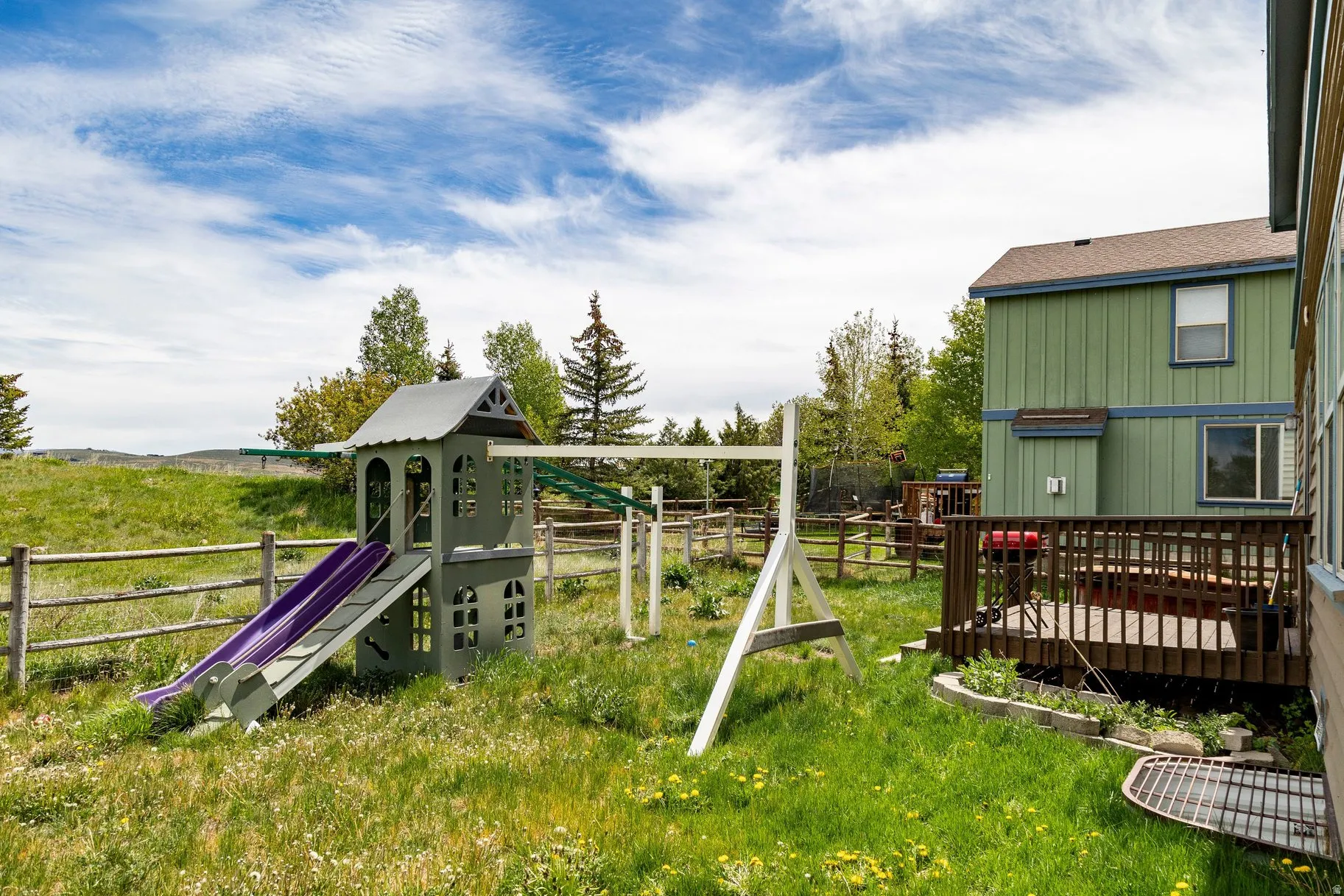 Expansive green space featuring a wooden split-rail fence, a raised wooden deck, and a two-story residential structure with green siding