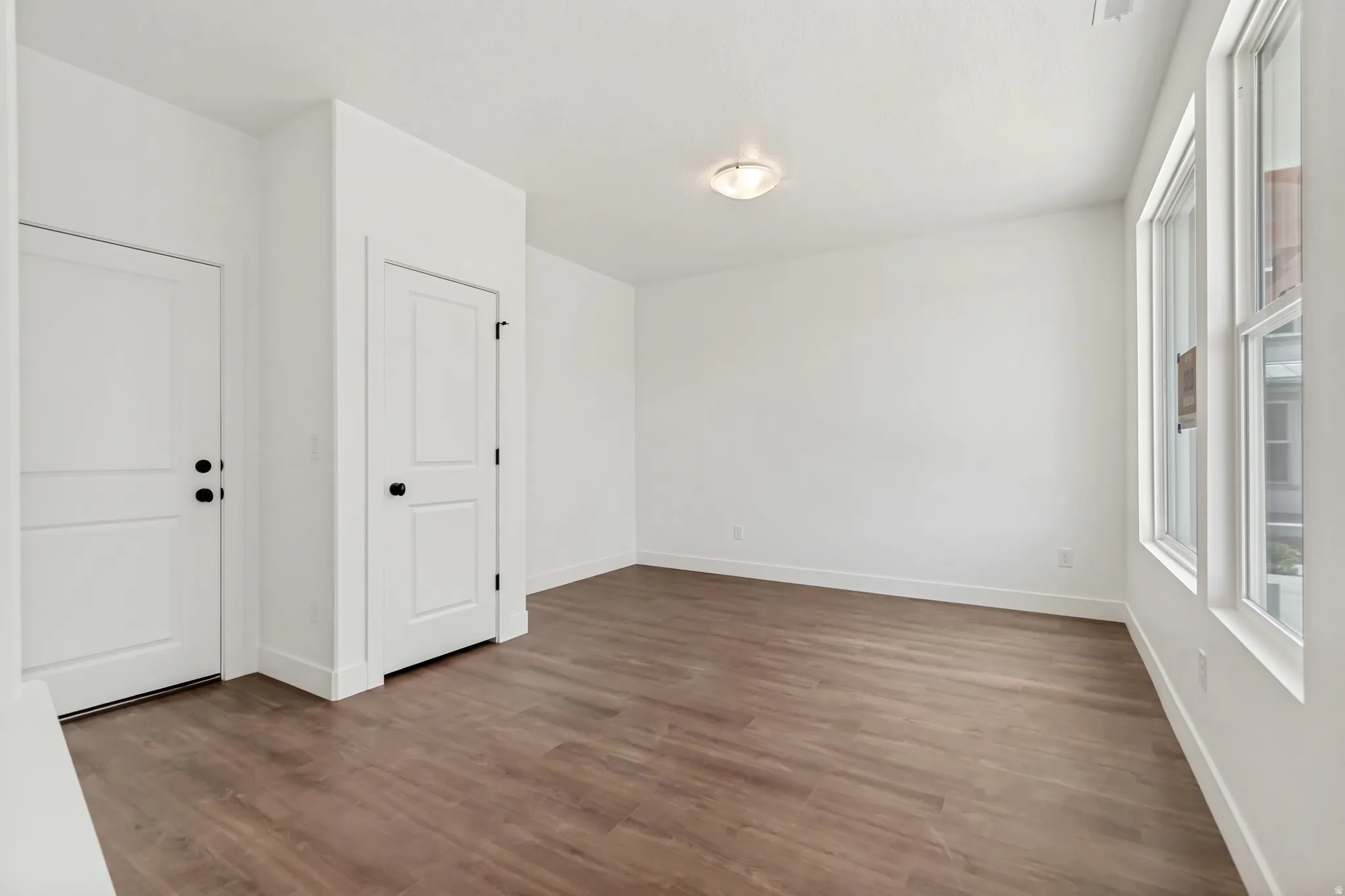 Bright room featuring wood-finish flooring, multiple windows, a ceiling-mounted light fixture, white walls, and two paneled doors with dark hardware