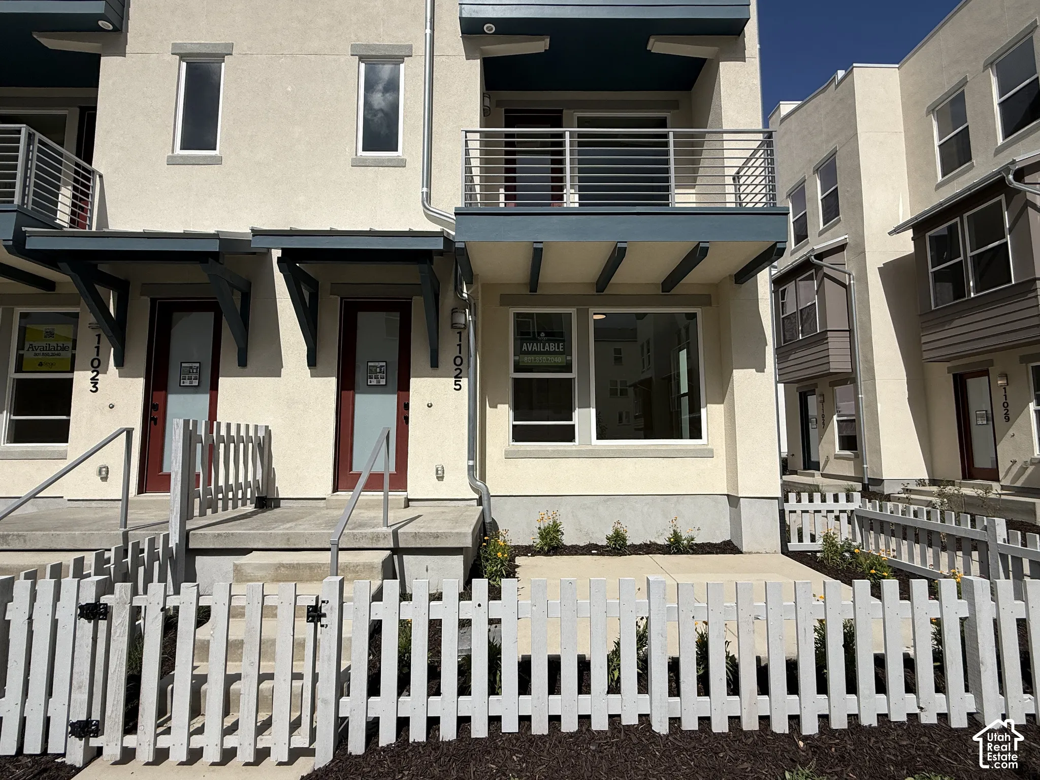 Stucco exterior with modern architectural elements, featuring a second-story balcony with metal railings, a covered entryway, and a white picket fence enclosing a landscaped front area
