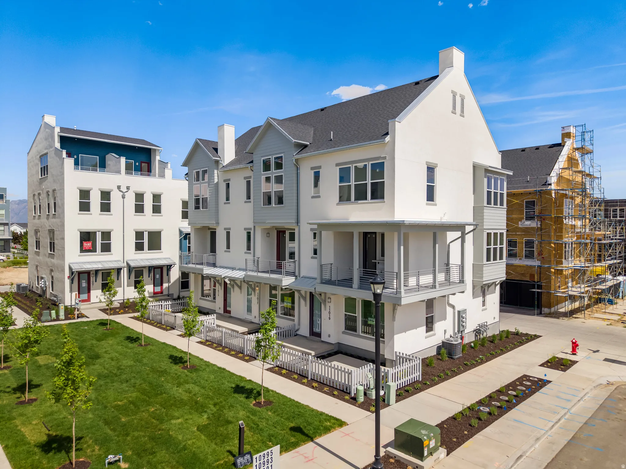 Modern townhouse exteriors featuring white and gray siding, dark shingle roofs, balconies with metal railings, and a landscaped green space with young trees