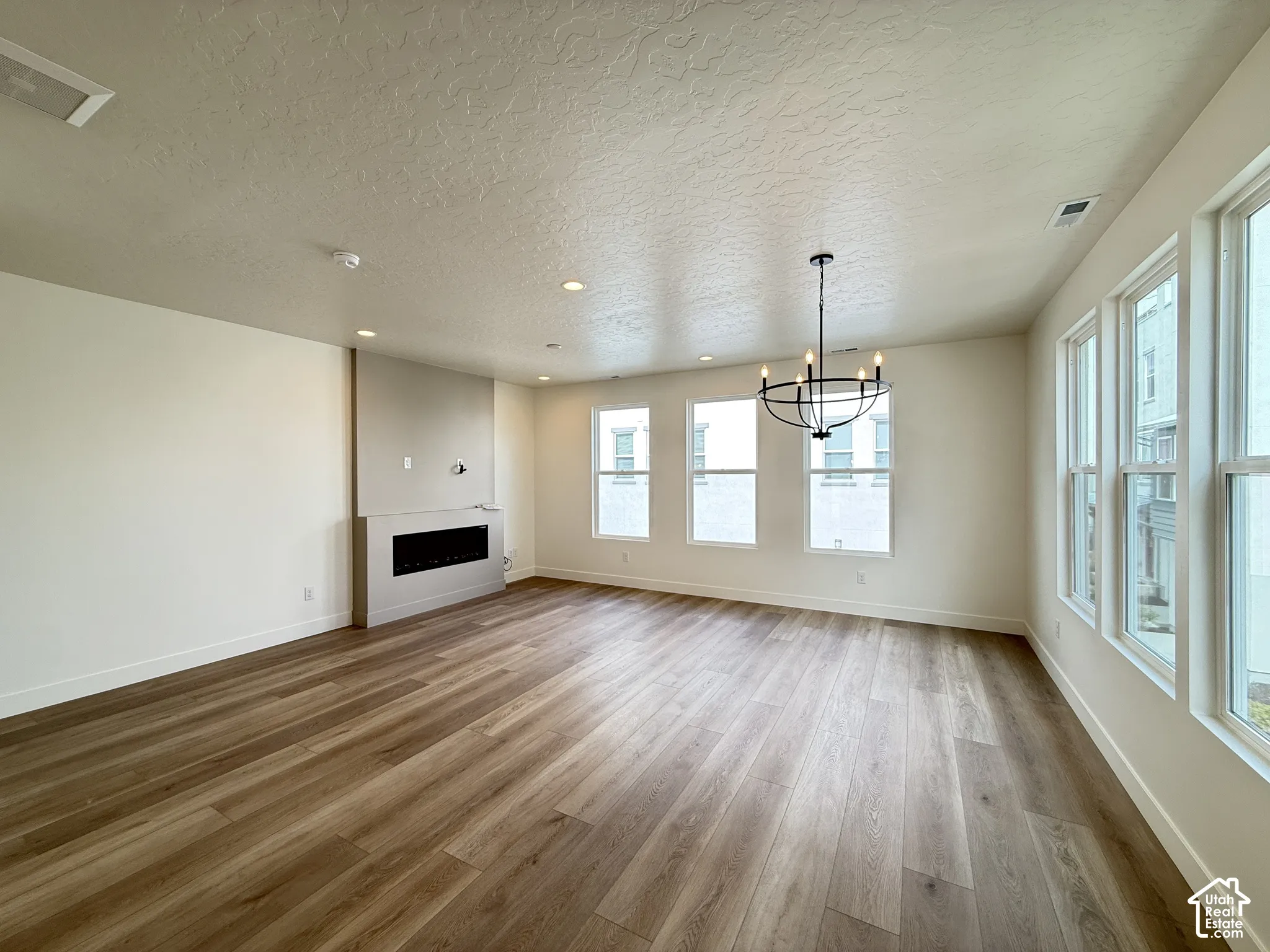 Spacious room featuring extensive wood-finish flooring, multiple large windows, a built-in electric fireplace, recessed lighting, and a contemporary chandelier
