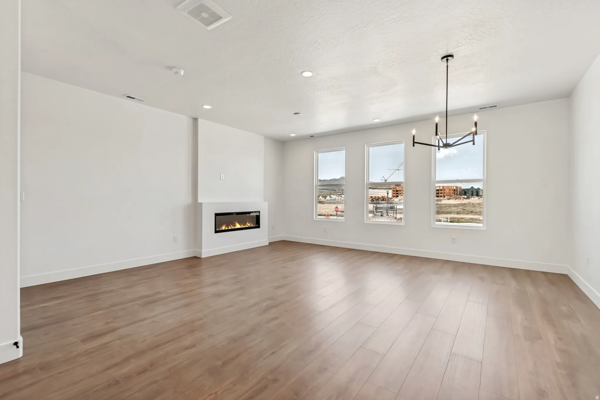 Spacious room featuring wood-finish flooring, an in-wall fireplace, three large windows, a contemporary black chandelier, and recessed lighting