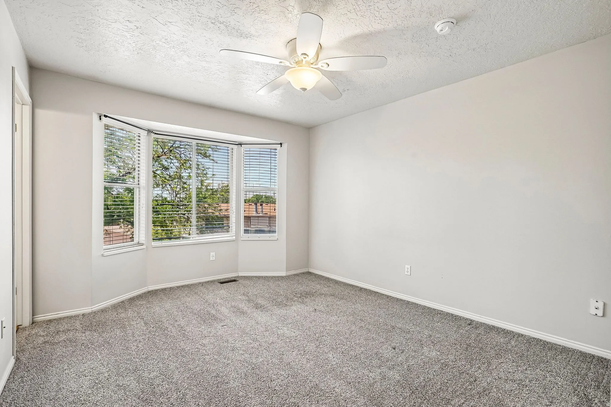 Spacious room featuring a bay window, neutral wall tones, gray carpeting, a ceiling fan, and white trim