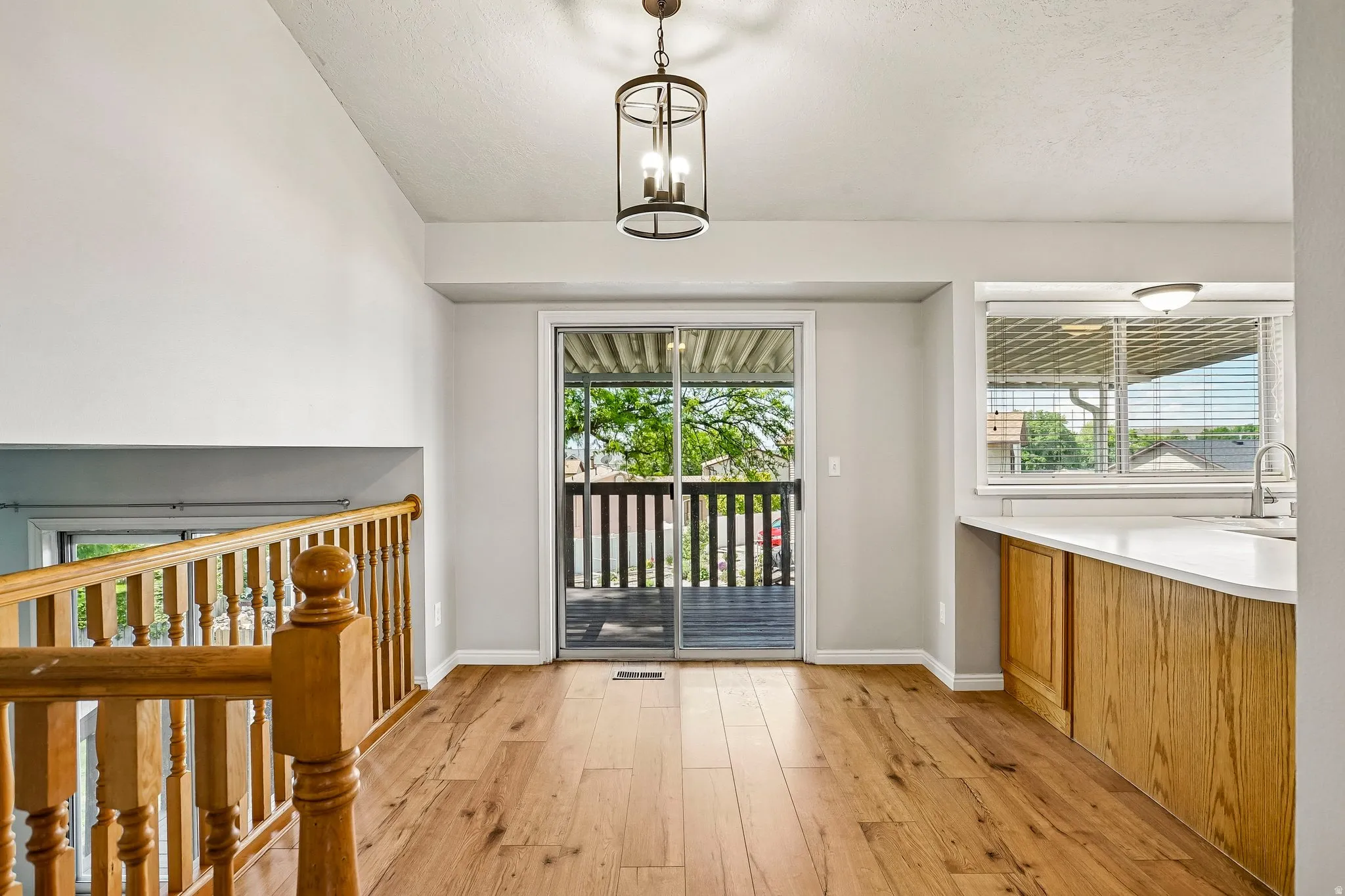 Wood-finish flooring throughout, featuring a modern chandelier, sliding glass door to an outdoor deck, and a kitchen peninsula with a white countertop and natural wood cabinetry