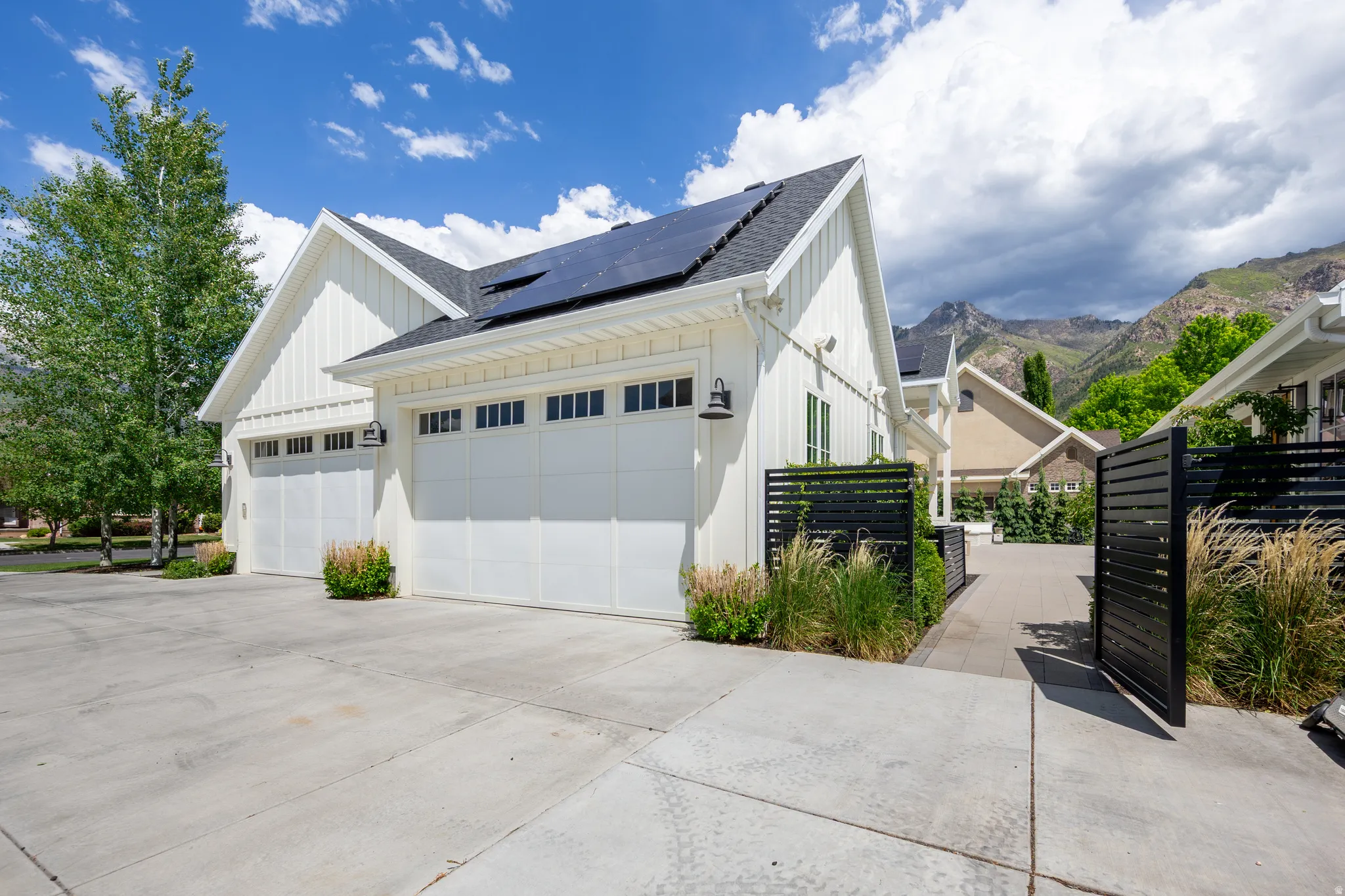 Modern farmhouse exterior with white board and batten siding, multiple garage doors with window accents, solar panels, a paved driveway, and black slatted fencing