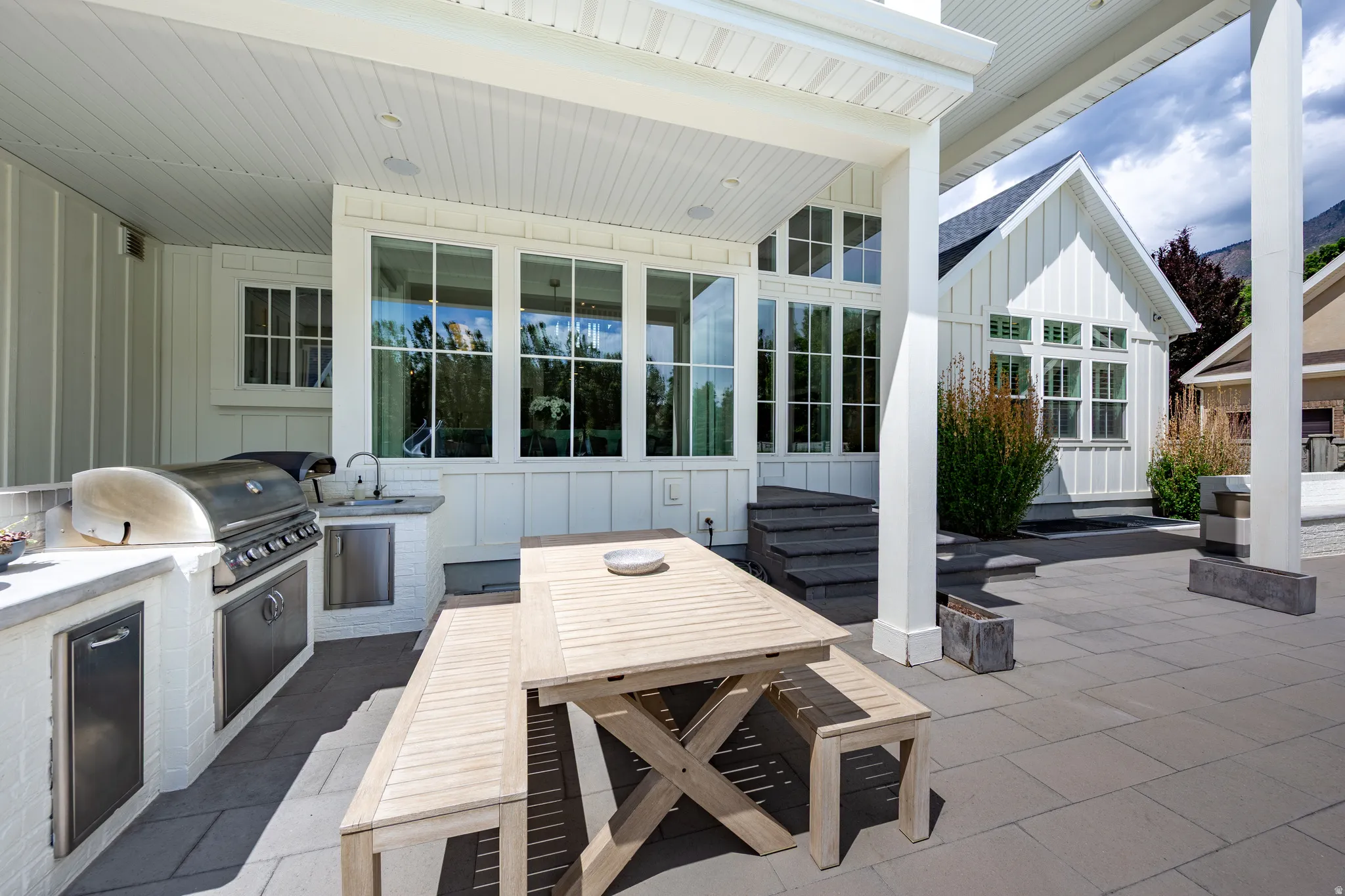 Expansive outdoor patio featuring an integrated outdoor kitchen with a stainless steel grill and sink, a covered dining area, and large format paver flooring