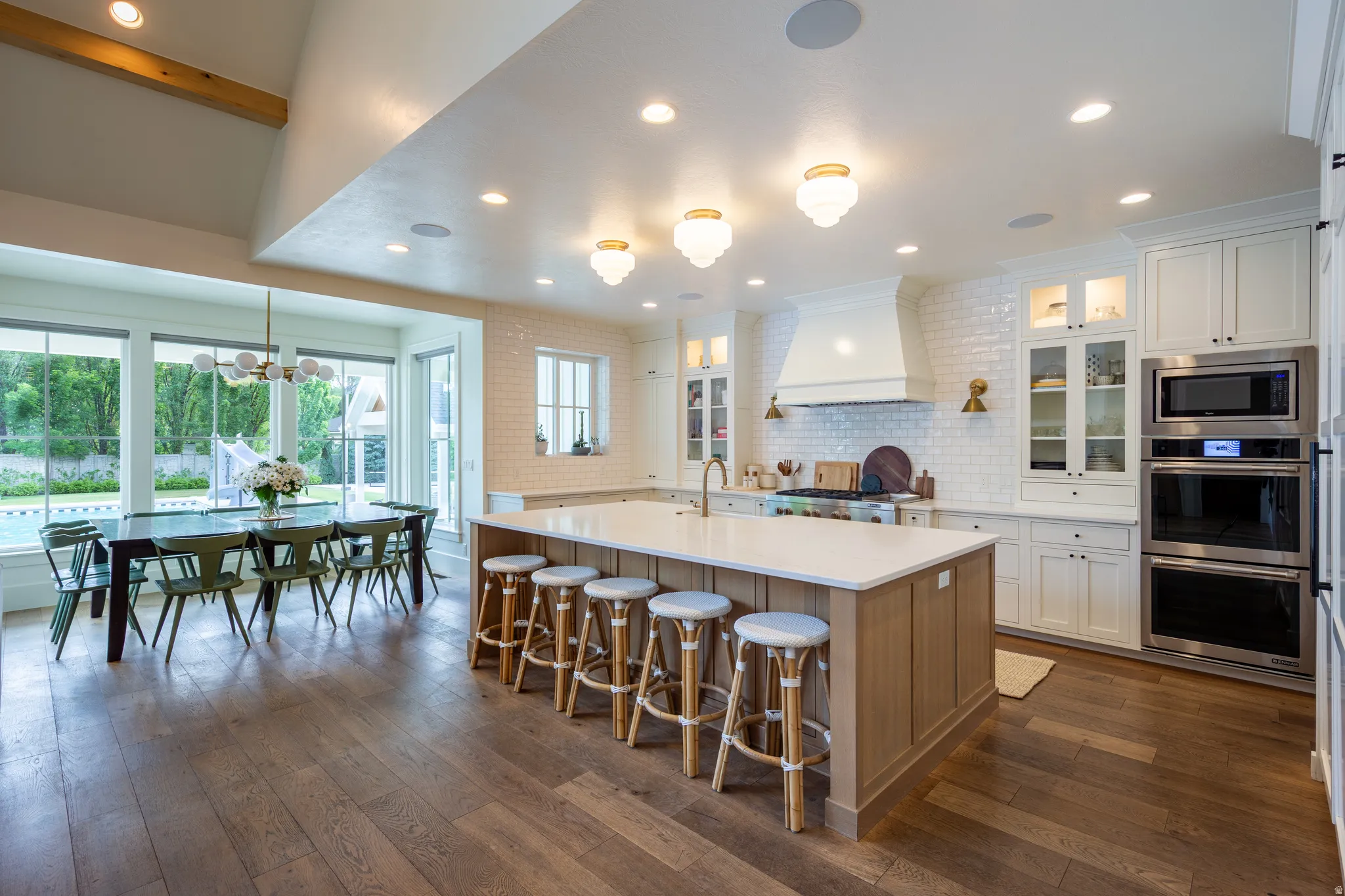 Gourmet kitchen with wood-finish flooring, a large center island with seating, integrated double ovens, and a dining area featuring full-height windows and a vaulted ceiling with an exposed beam