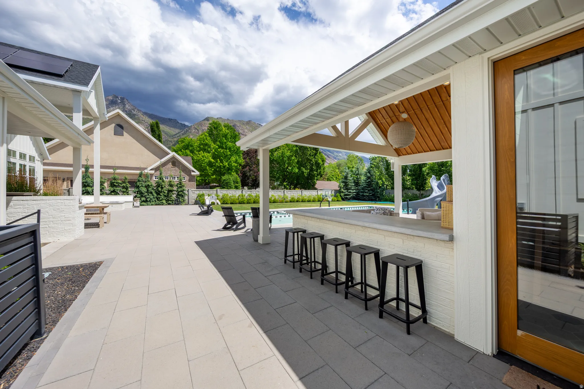 Expansive outdoor patio with large format pavers, a covered bar area featuring a wood-finish ceiling, an outdoor pool, mature landscaping, and mountain scenery