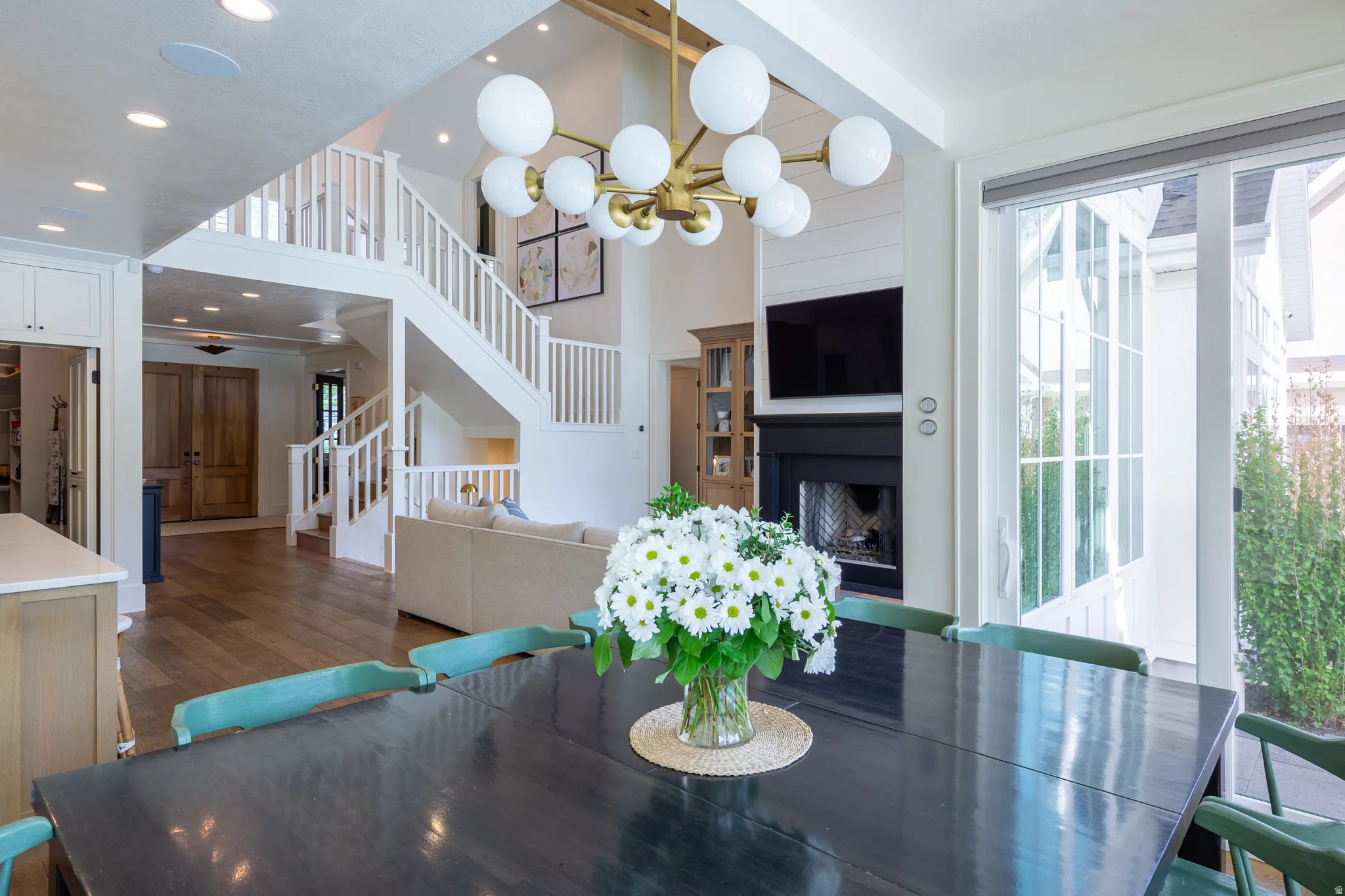 Open-concept living area featuring wood-finish flooring, a contemporary chandelier, a two-story staircase with white railings, a fireplace with shiplap detailing, and large glass sliding doors