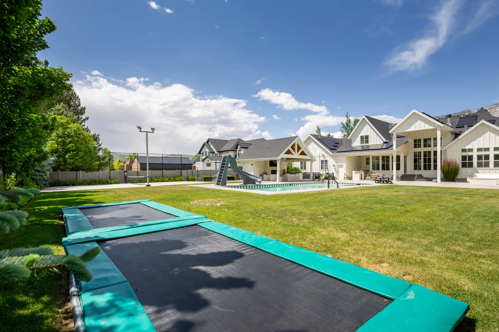 Expansive rear yard featuring a manicured lawn, inground trampolines, and a dedicated sports court with a light pole