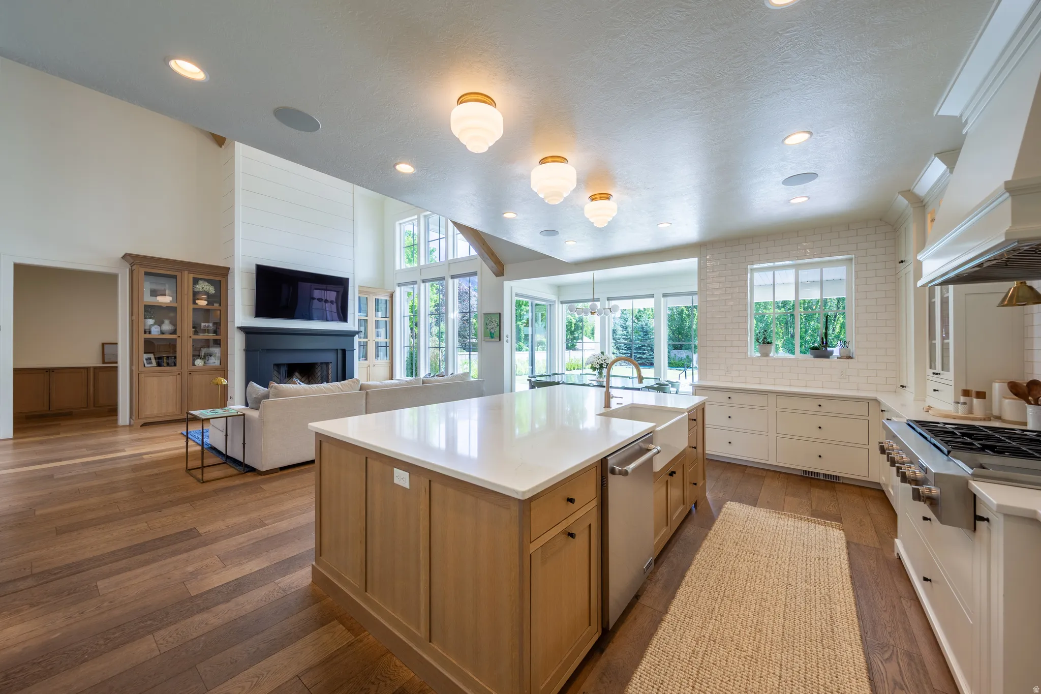 Open-concept kitchen and living area featuring wood-finish flooring, a spacious kitchen island with a farmhouse sink, subway tile backsplash, a professional-grade range with a custom hood, and abundant natural light from large windows and glass doors