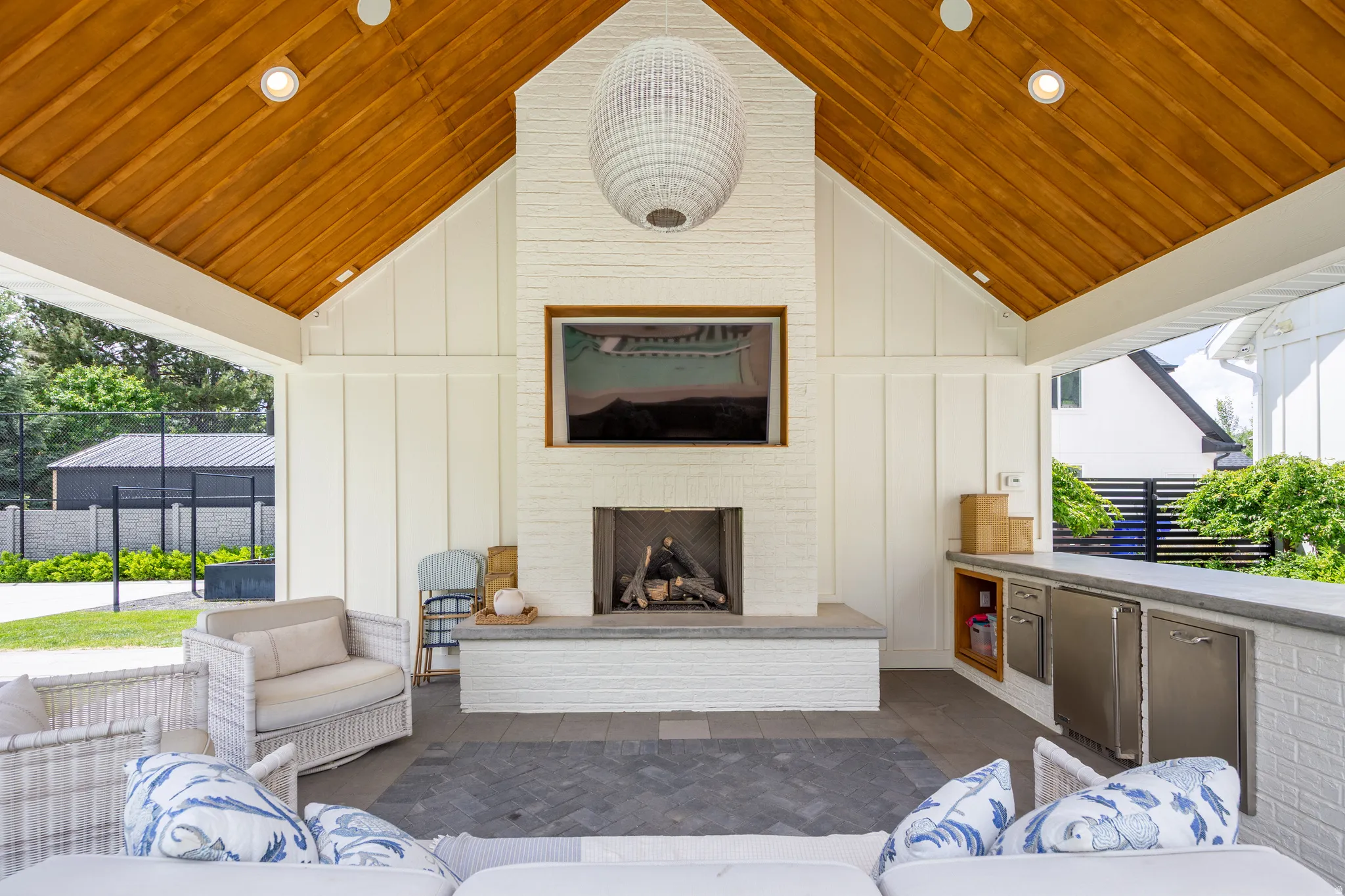 Outdoor entertaining space featuring a vaulted wood-plank ceiling, white board-and-batten walls, a white brick fireplace with a stone hearth, and integrated outdoor kitchen appliances