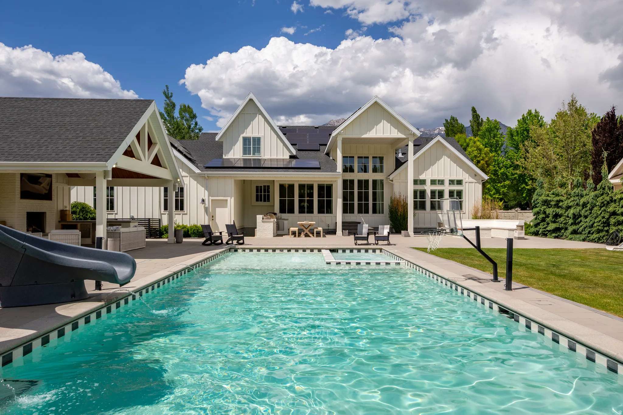 Spacious backyard featuring a rectangular swimming pool with a slide, extensive concrete patio, and a covered outdoor living area with a built-in fireplace
