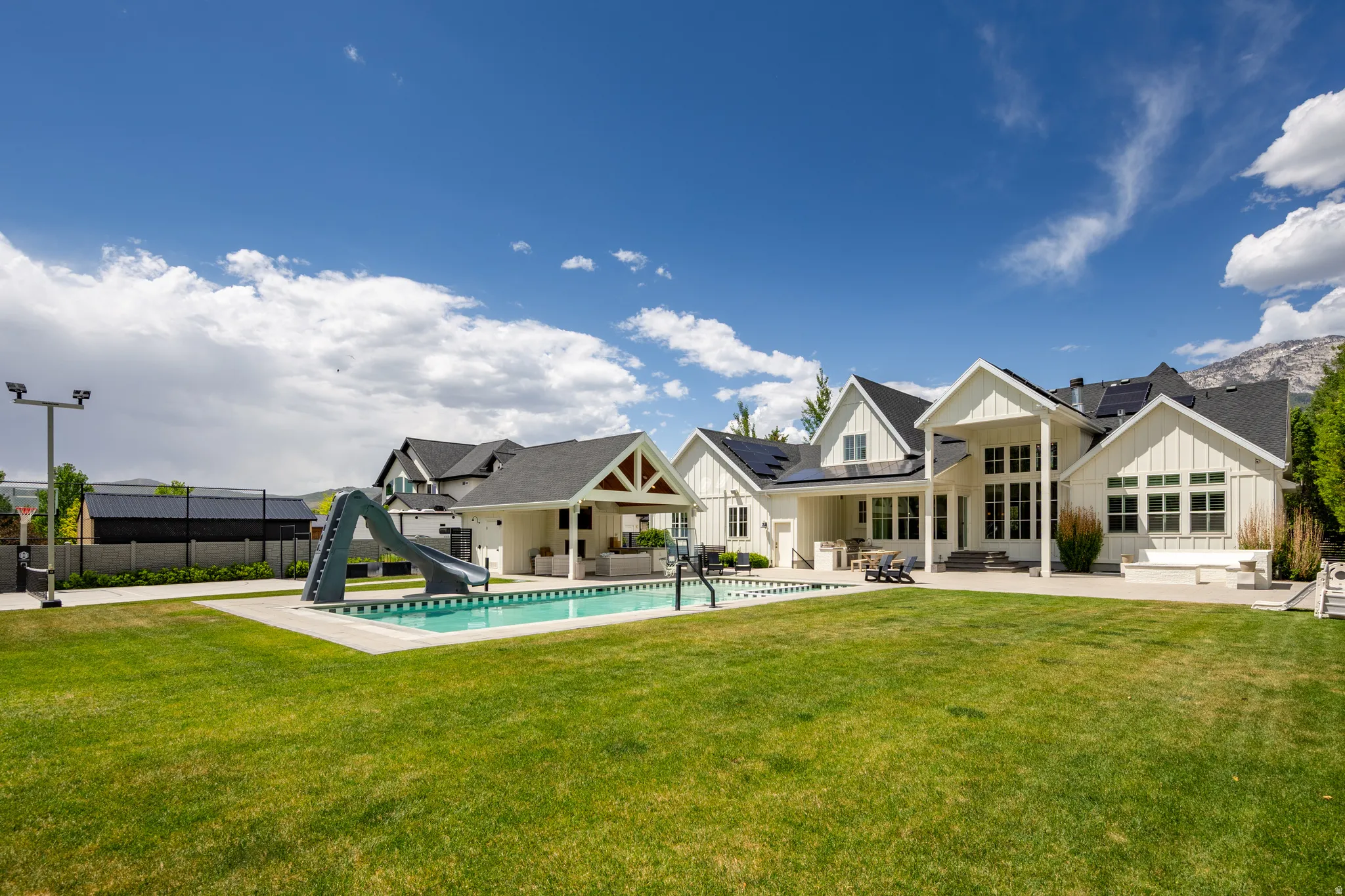 Expansive backyard featuring a rectangular swimming pool with integrated slide, a covered patio area, a concrete sport court, and a vast lawn