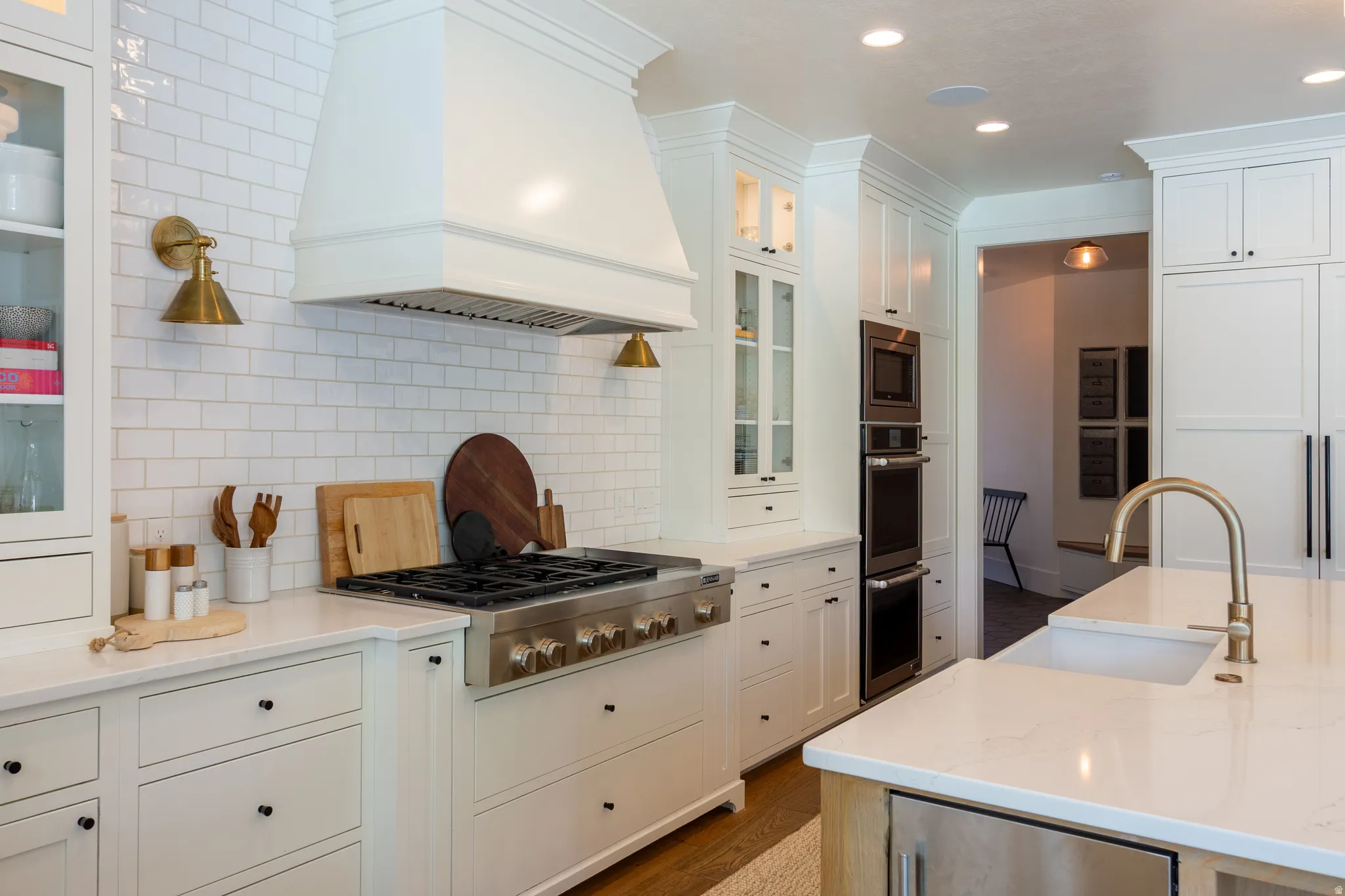 Gourmet kitchen featuring a professional-grade stainless steel range, custom white range hood, white subway tile backsplash, built-in double oven, and a large island with an undermount sink and brass faucet