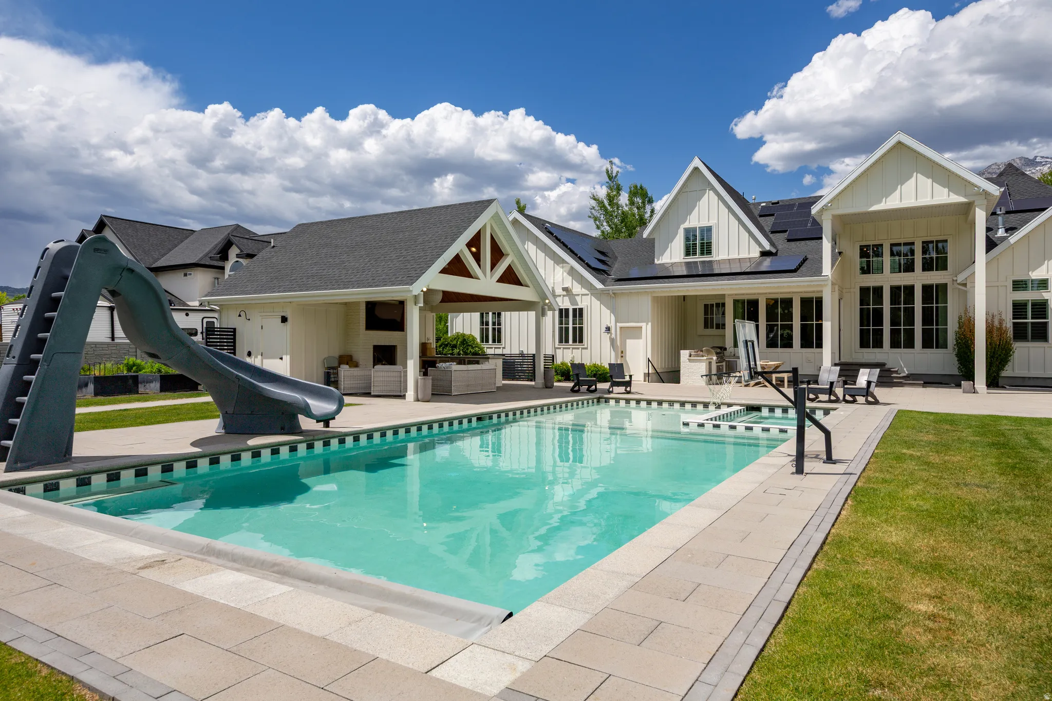 Expansive outdoor living area featuring a rectangular swimming pool with integrated spa and pool slide, complemented by a covered patio with exposed beam detailing and an outdoor fireplace