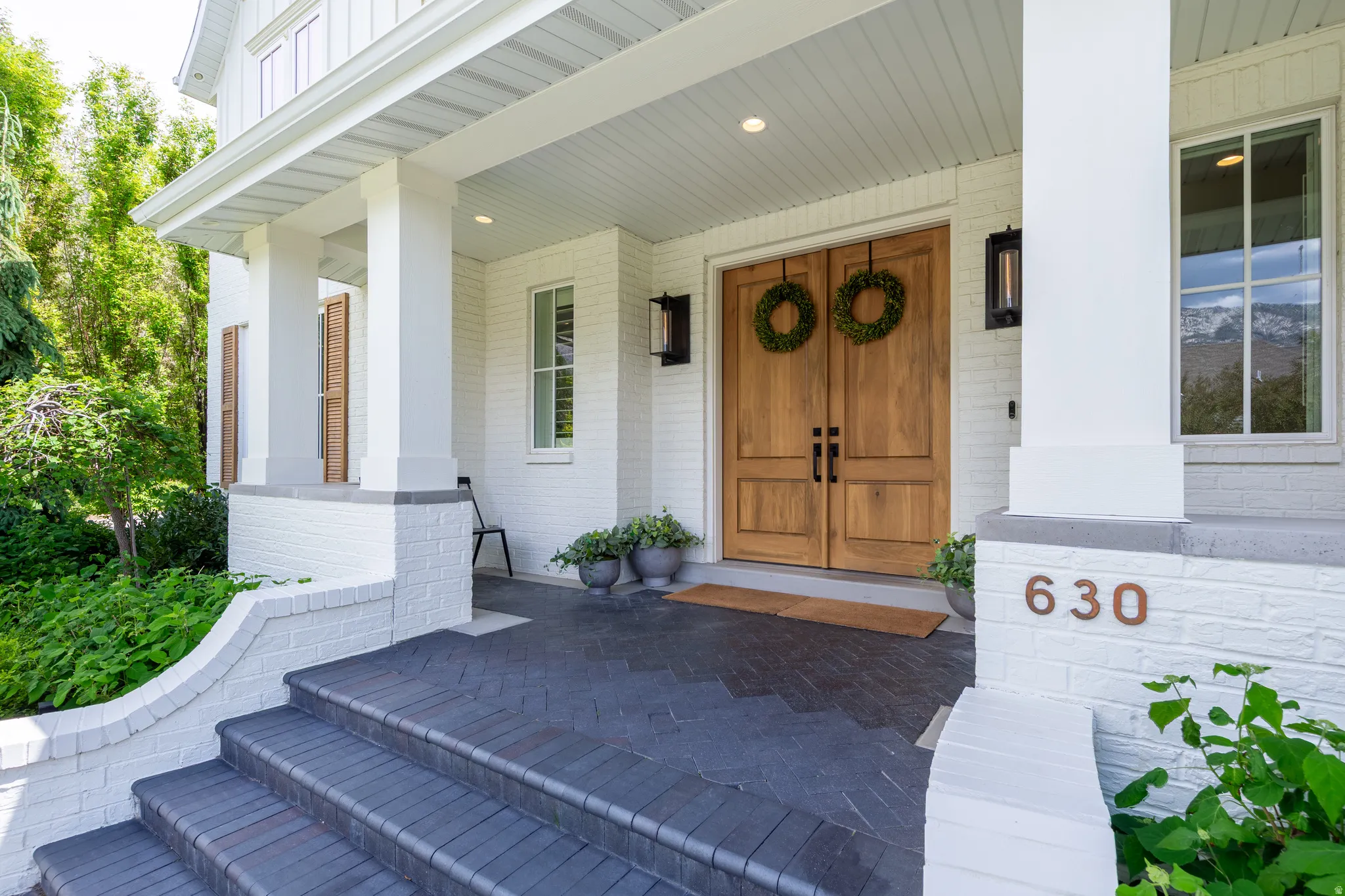 Inviting front entrance featuring a covered porch, white painted brick facade, natural wood double entry doors, dark-toned brick stairs, and rectangular outdoor sconces