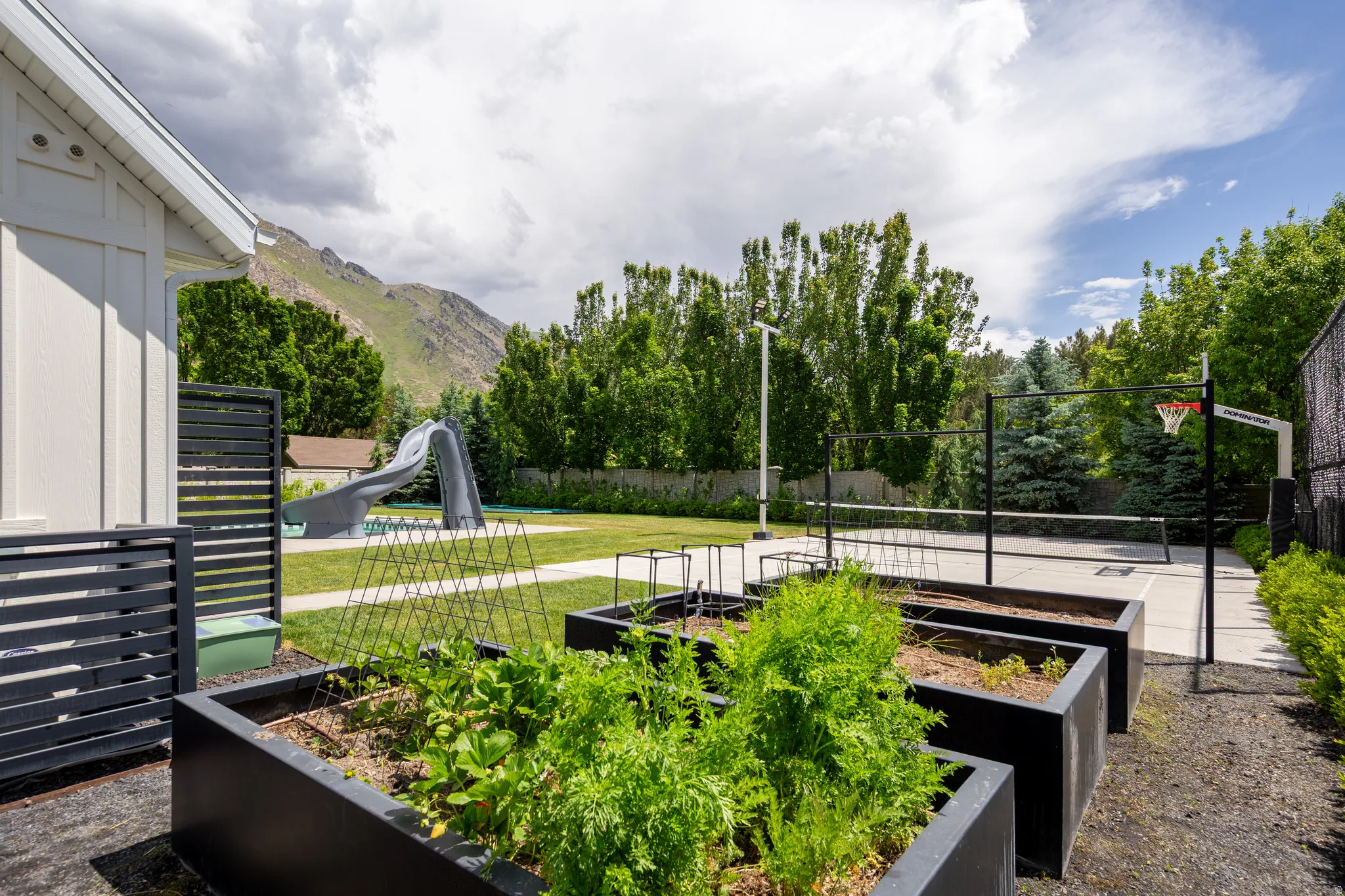 Expansive backyard featuring a manicured lawn, an in-ground swimming pool with a slide, a multi-sport court with a basketball hoop and net, and raised garden beds