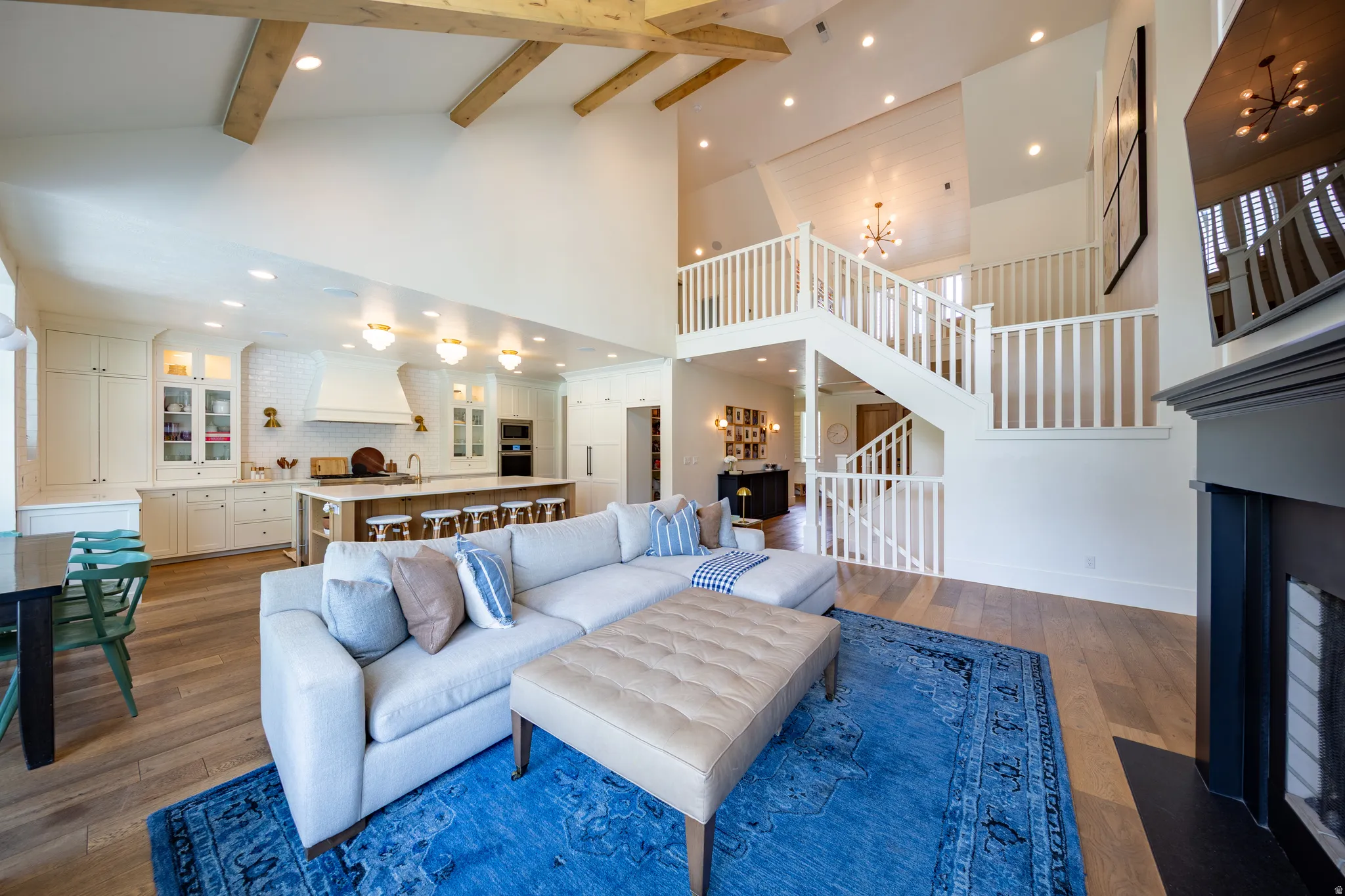 Vaulted ceiling with exposed wood beams, wood-finish flooring, built-in kitchen cabinetry with glass panel accents, multi-level staircase with white railings, and a fireplace with a dark mantel