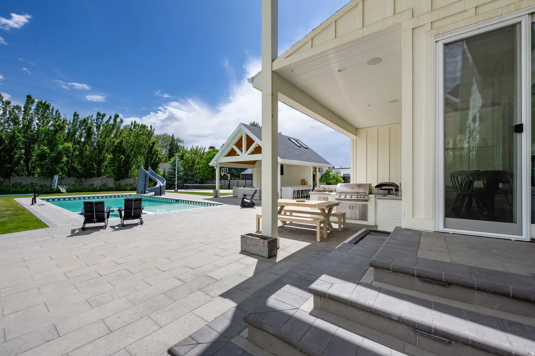 Expansive outdoor living area featuring a rectangular pool with a waterslide, a large paved patio, a covered outdoor kitchen with built-in grill, a separate covered structure, and mature perimeter trees