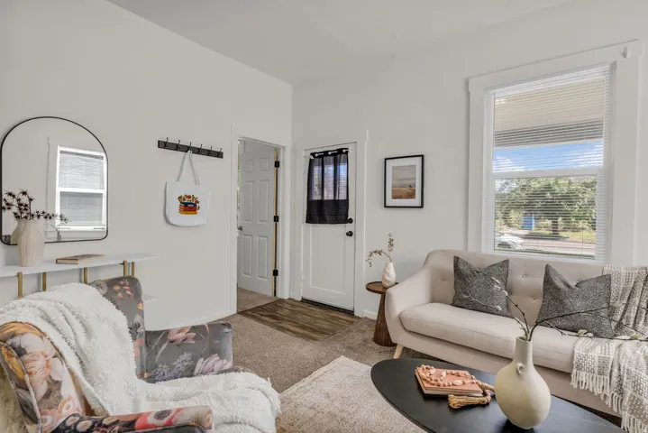 Living space featuring neutral wall tones, carpeted flooring, a white trim window, a white paneled interior door, and a partially visible wood-finish flooring entryway