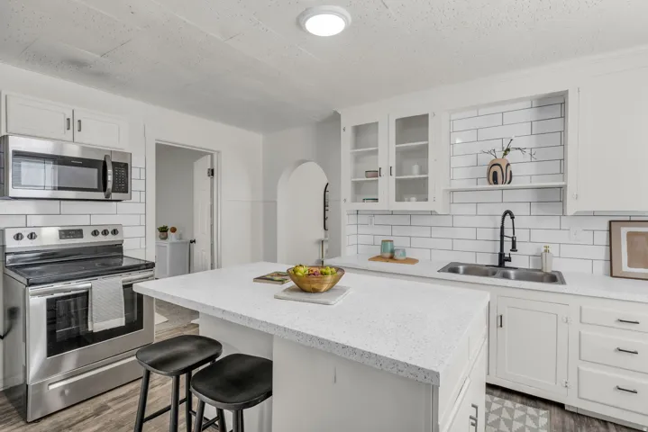 Modern kitchen featuring a central island with a light-colored countertop, stainless steel appliances, a subway tile backsplash, white cabinetry with glass-front uppers, and wood-finish flooring