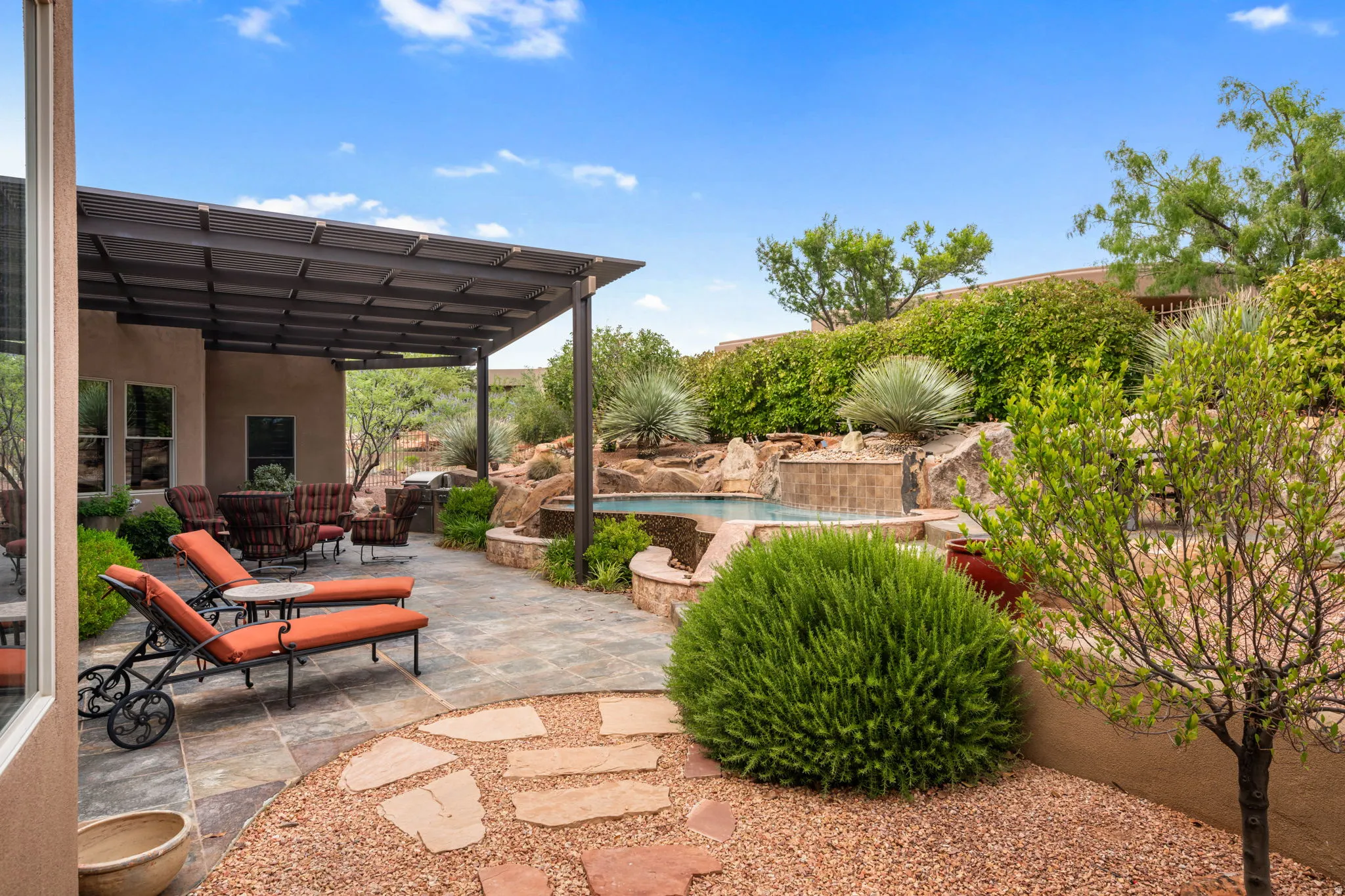 Expansive outdoor living area featuring a covered patio with a dark metal pergola, flagstone paving, a built-in pool with rock-stack surround and waterfall feature, and established landscaping