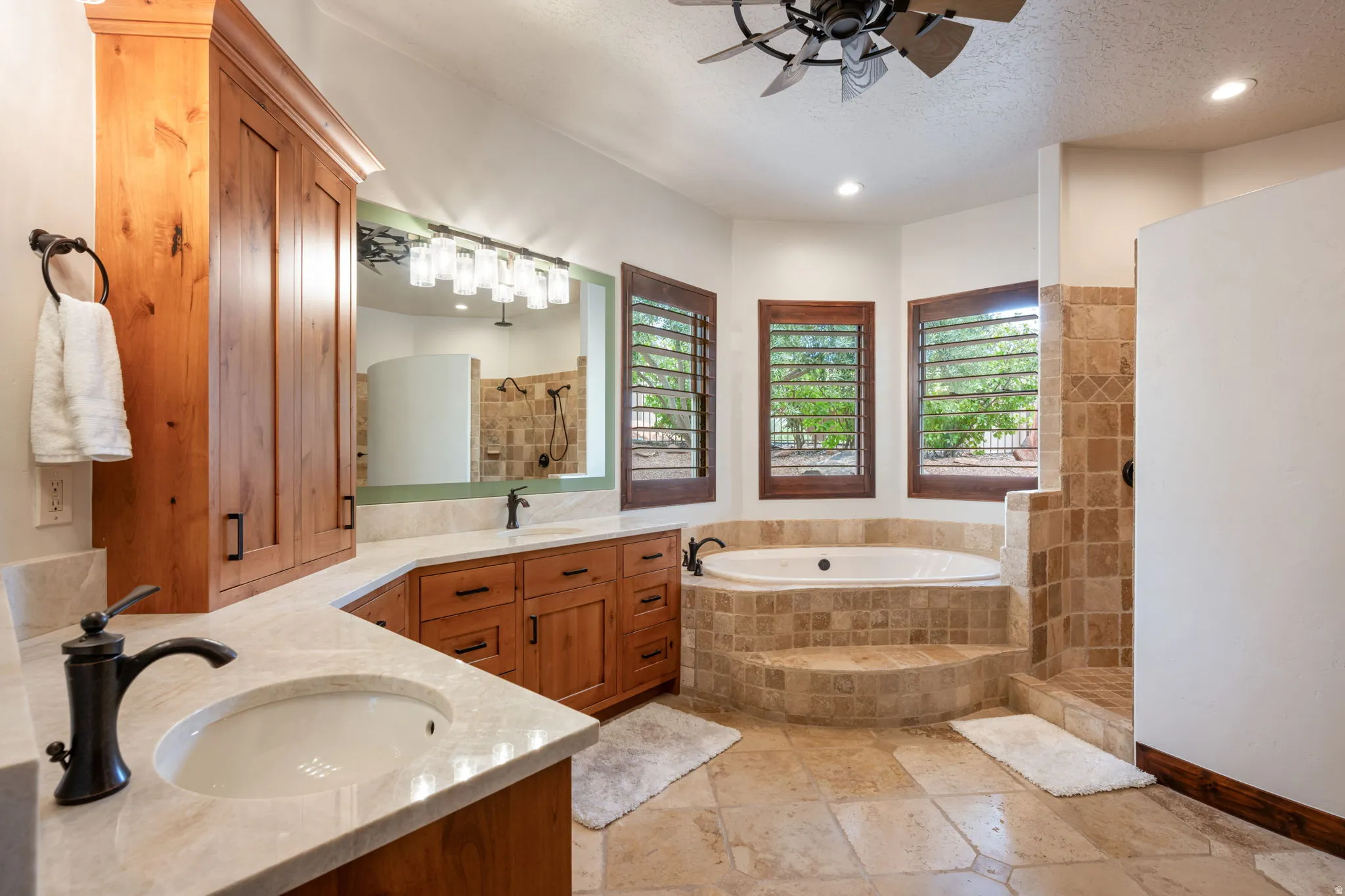 Spacious bathroom featuring a built-in soaking tub with stone tile surround, a dual vanity with light-toned countertops and integrated sinks, natural wood cabinetry, and three windows with wooden plantation shutters