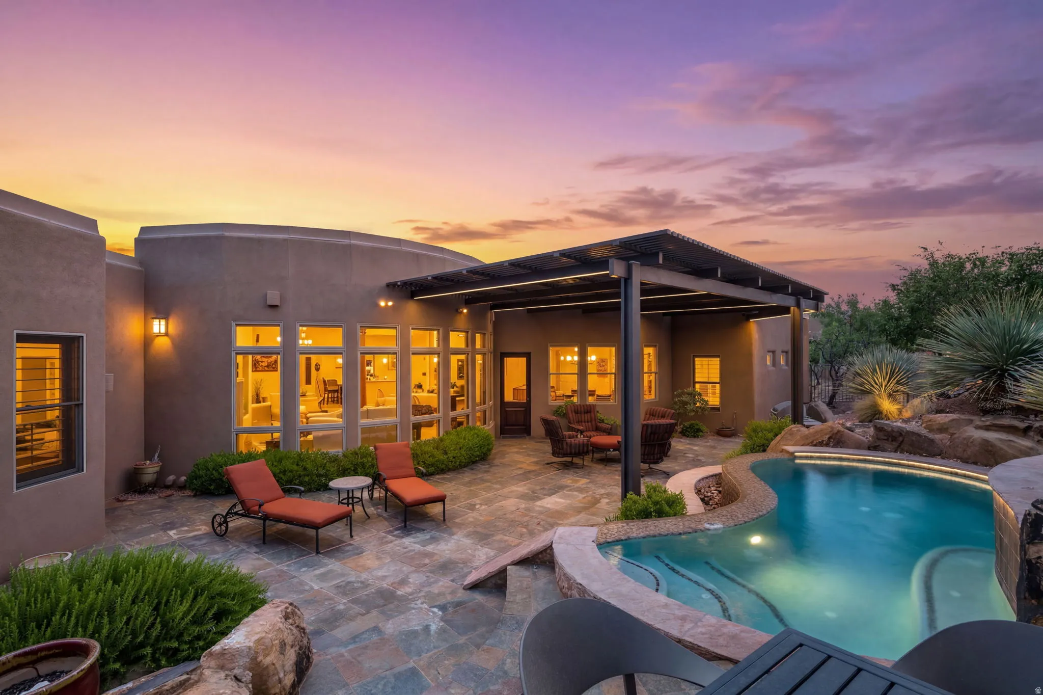 Curved stucco exterior with a large patio featuring multi-toned flagstone, a covered outdoor living space with a slatted roof, an irregularly shaped swimming pool, and desert landscaping with accent lighting