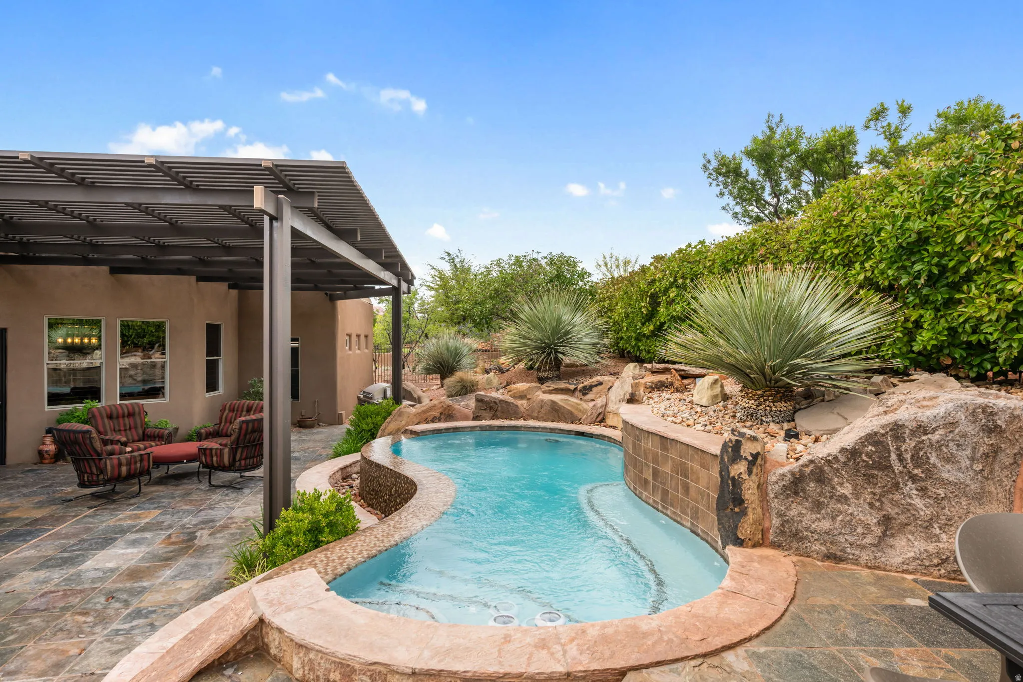 Curved swimming pool with integrated spa and rock waterfall feature, complemented by a covered patio with an open slat pergola