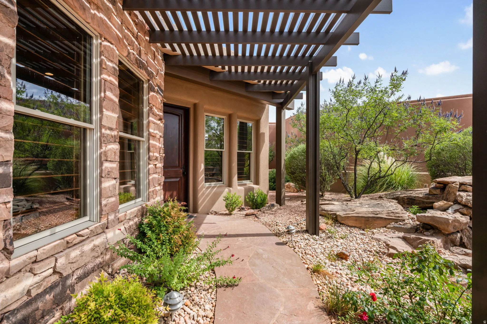 Stone veneer exterior with multiple windows, a dark wood entry door, a slatted patio cover, a flagstone walkway, and desert landscaping featuring natural rock elements
