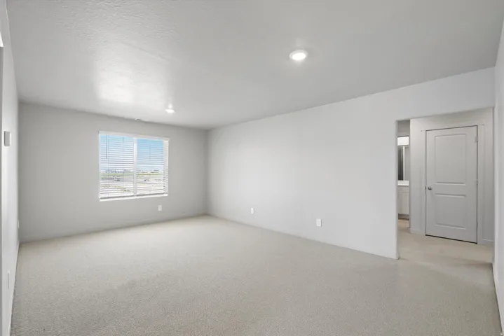 Spacious room featuring light-colored carpeting, a large window with blinds, recessed ceiling lighting, and a white paneled door