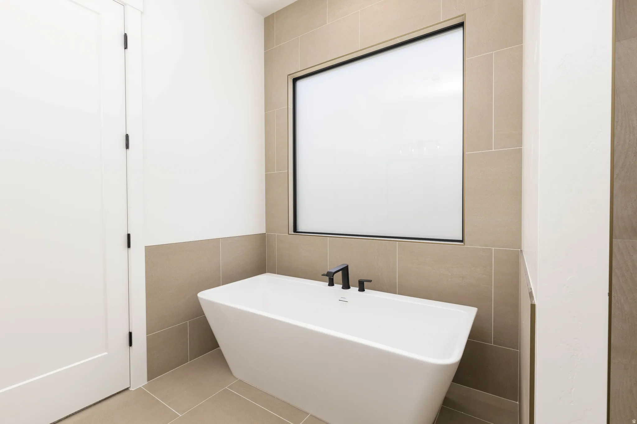 Freestanding soaking tub with matte black fixtures, set against a tiled wall with an integrated frosted window