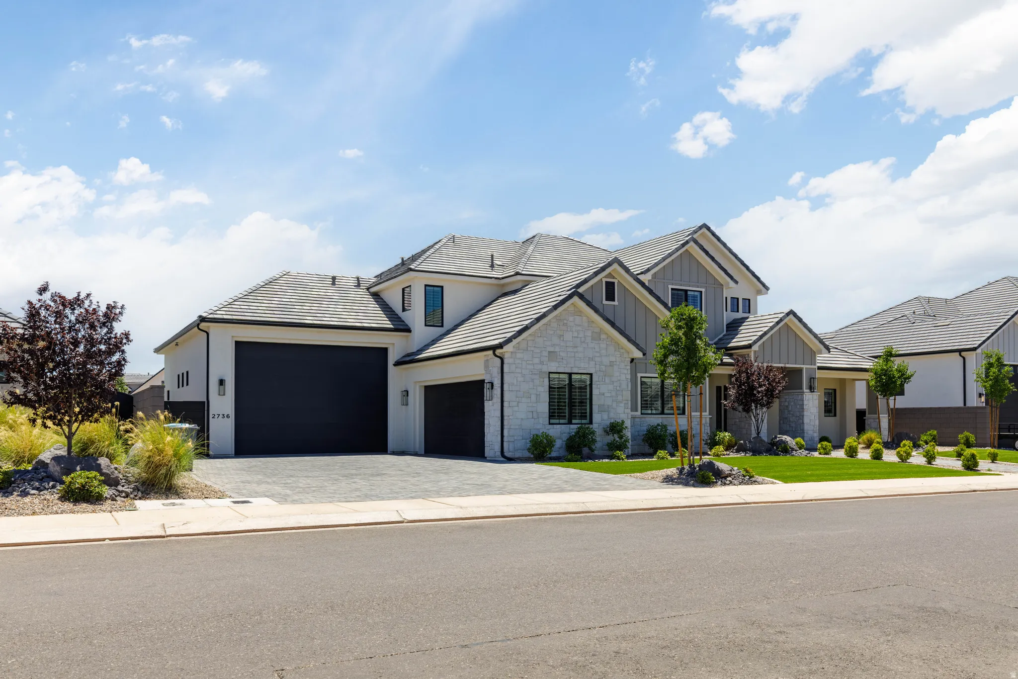 Contemporary residence featuring a light-colored stucco and stone exterior, dark garage doors, a paver driveway, pitched tile roof, and manicured landscaping