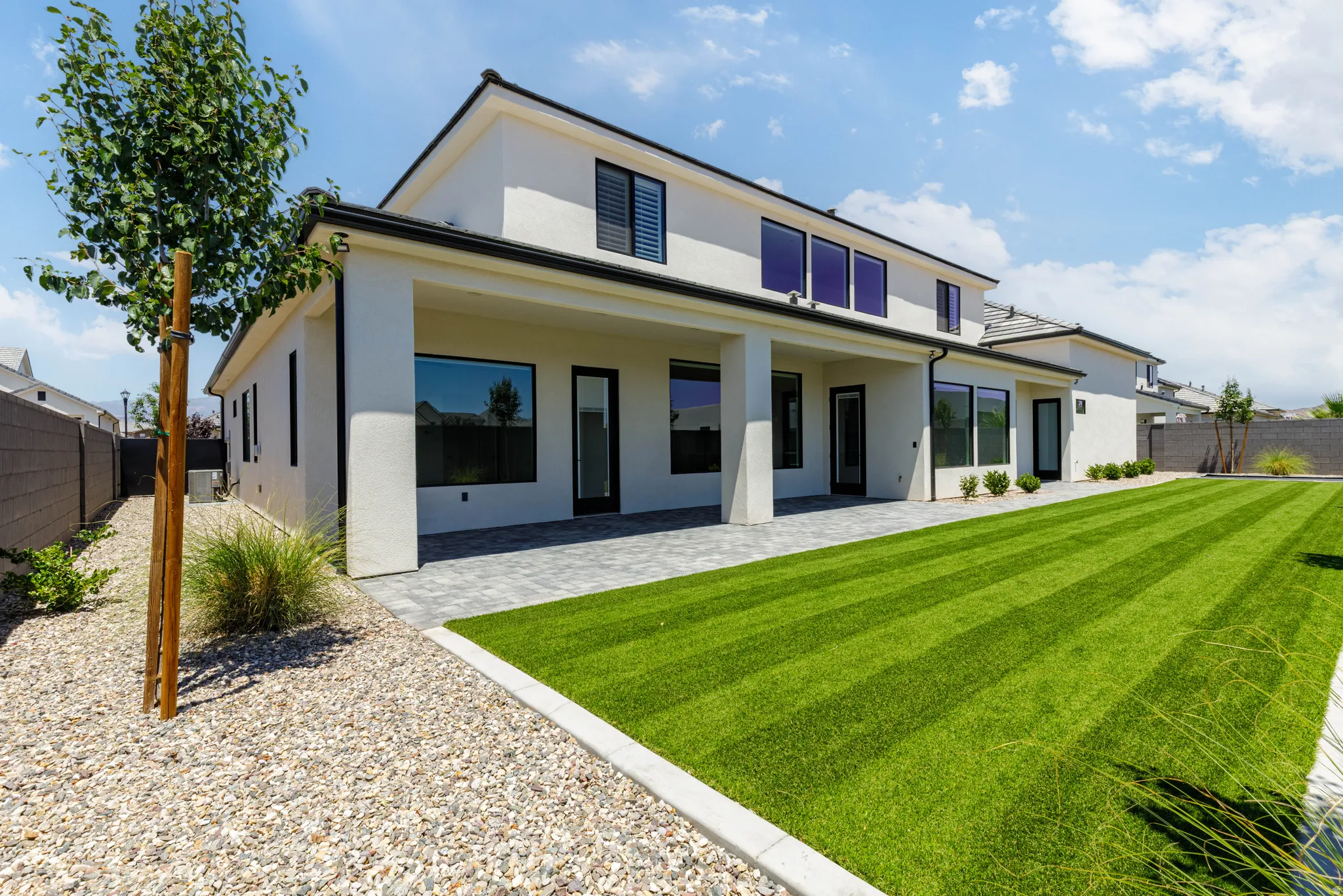 Rear exterior featuring a covered patio with paver flooring, expansive artificial turf lawn, xeriscape landscaping, and a stucco facade with black-framed windows