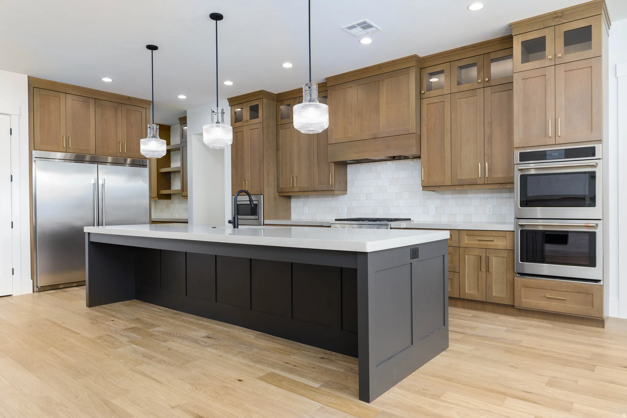 Gourmet kitchen featuring a large central island with a light-colored countertop, light wood-finish cabinetry, a white subway tile backsplash, and stainless steel appliances including a double oven and refrigerator