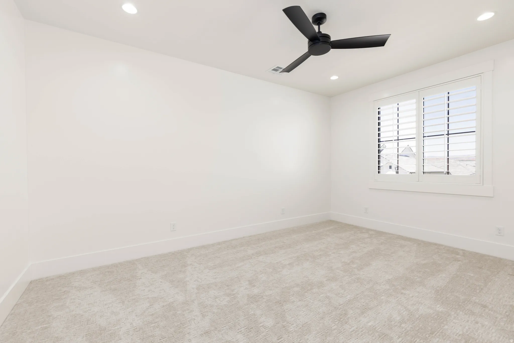 Spacious room featuring neutral carpeting, white walls, and a window with white plantation shutters