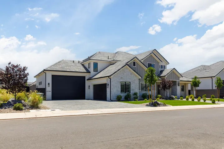Contemporary residence featuring a light-colored stucco and stone exterior, dark garage doors, a paver driveway, pitched tile roof, and manicured landscaping