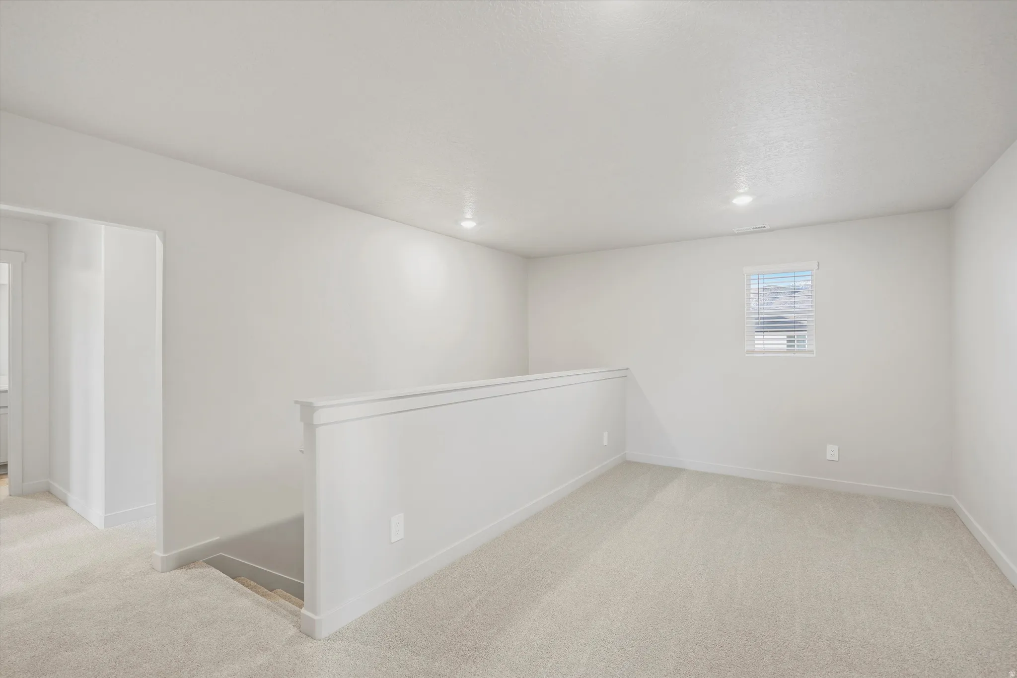 Carpeted upper level lounge with a window featuring horizontal blinds, recessed lighting, and a white wall-mounted railing overlooking a lower level