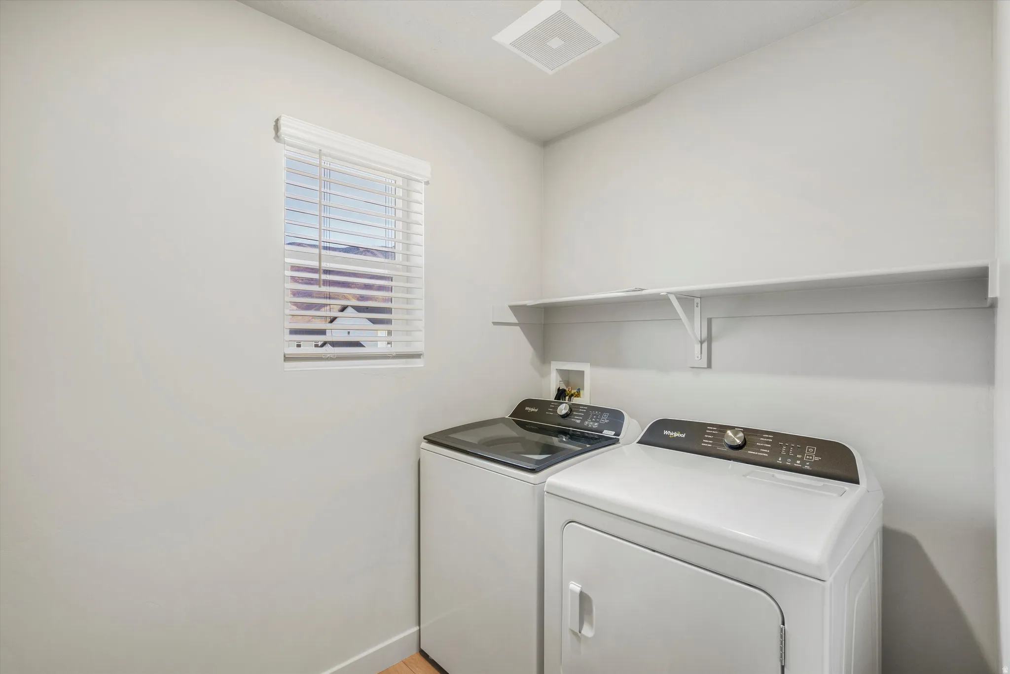 Dedicated laundry area featuring a window with white blinds, overhead shelving, and light wood-finish flooring