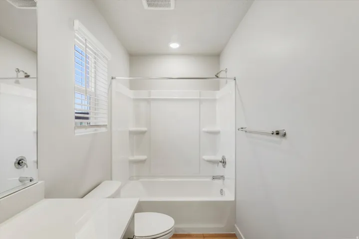 Bathroom featuring a white tub-shower combination unit, white toilet, and a window with white blinds