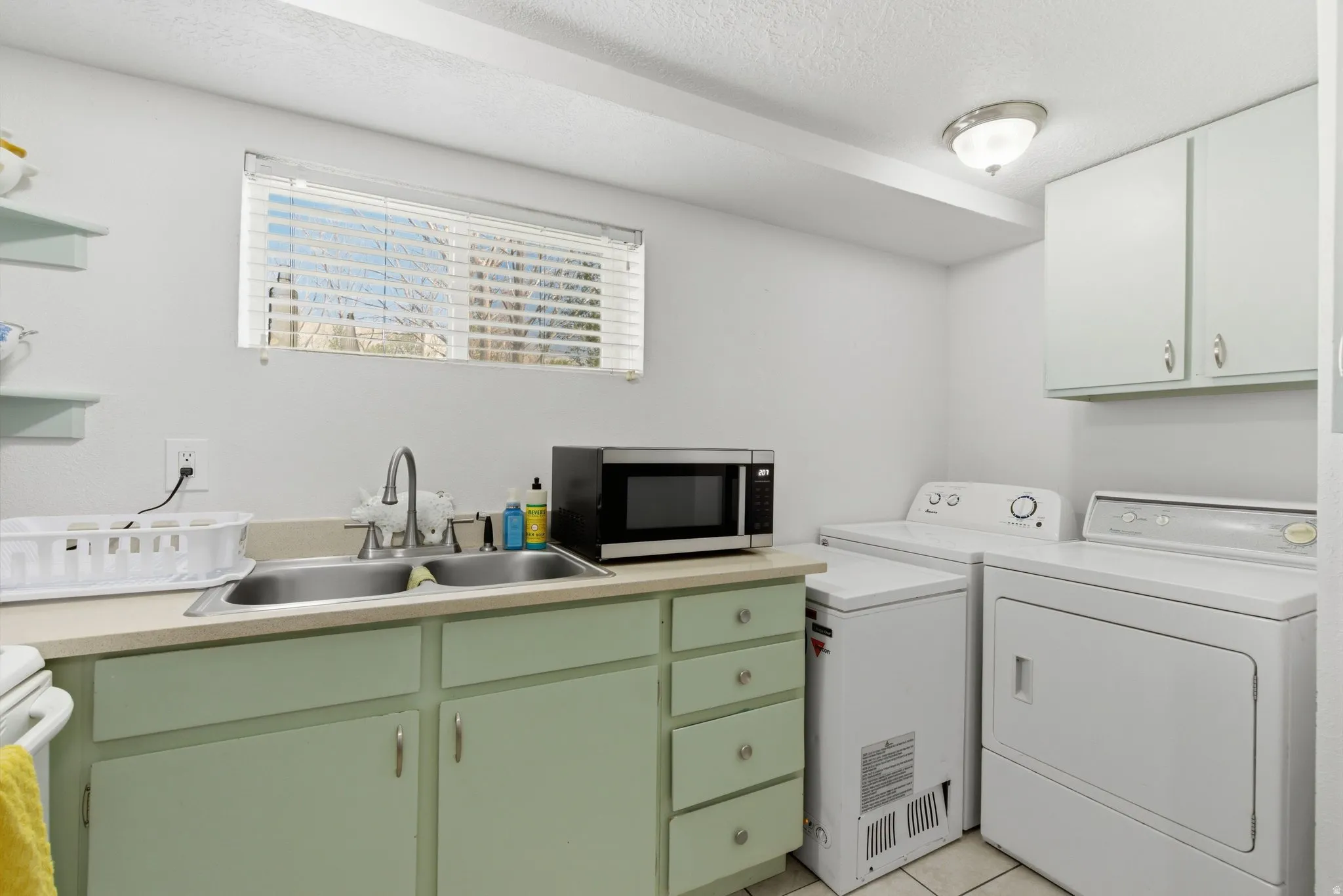 Utility room featuring a double basin stainless steel sink, light green cabinetry, upper white cabinetry, a window with blinds, and tiled flooring