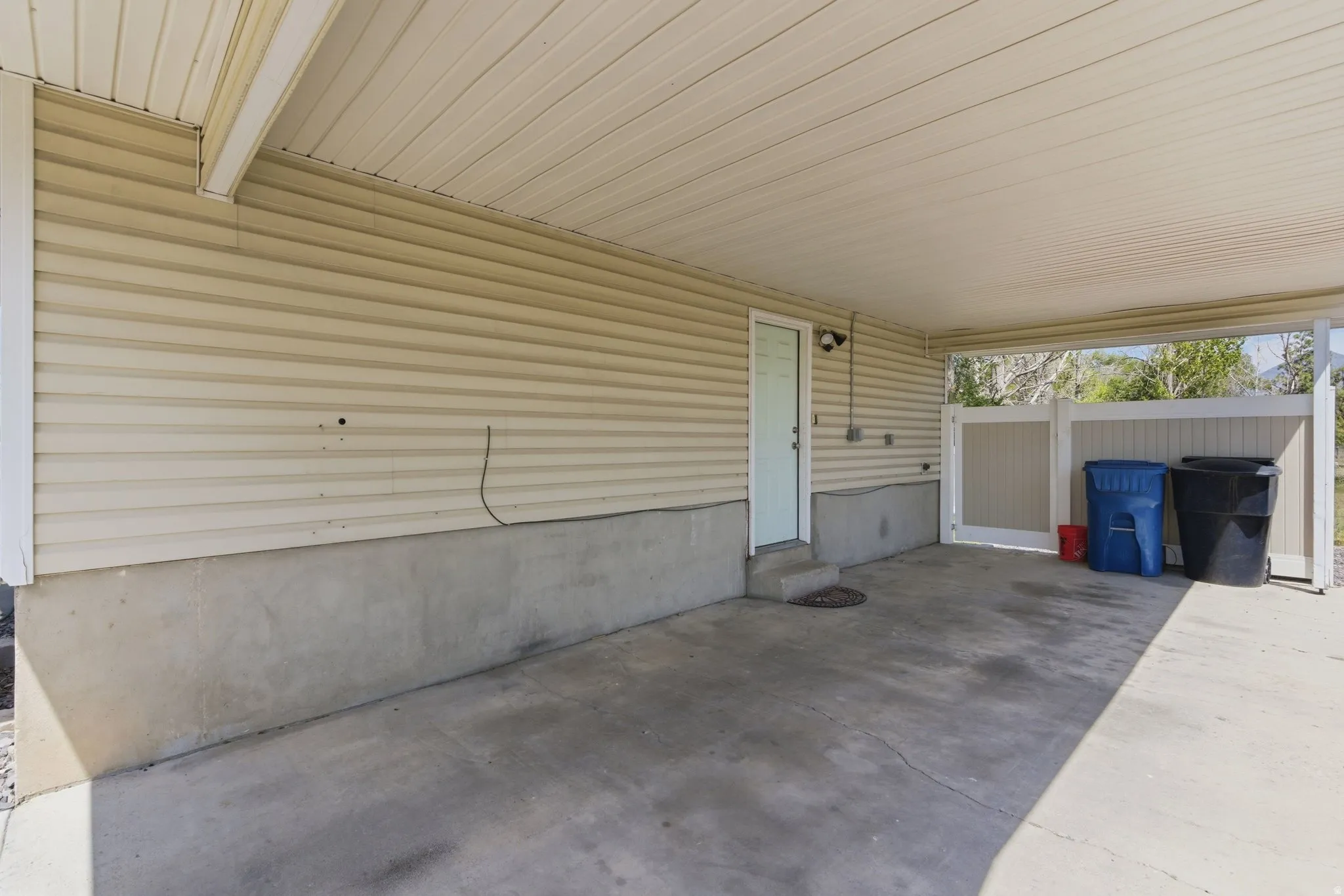 Carport with a concrete floor, vinyl siding, a white service door, and a privacy fence