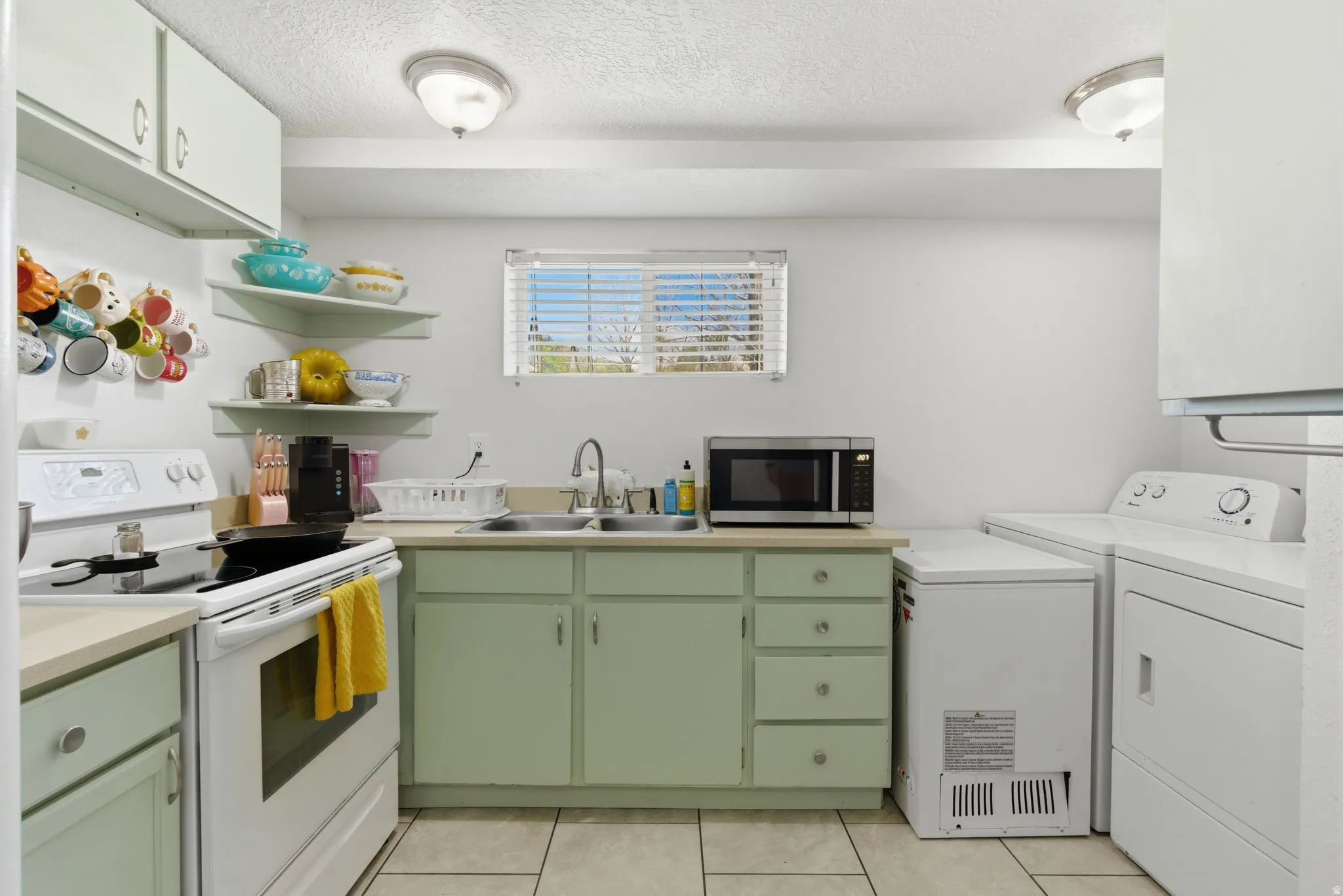 Kitchenette featuring light green cabinetry, white appliances, tiled flooring, a stainless steel sink, and a window with blinds