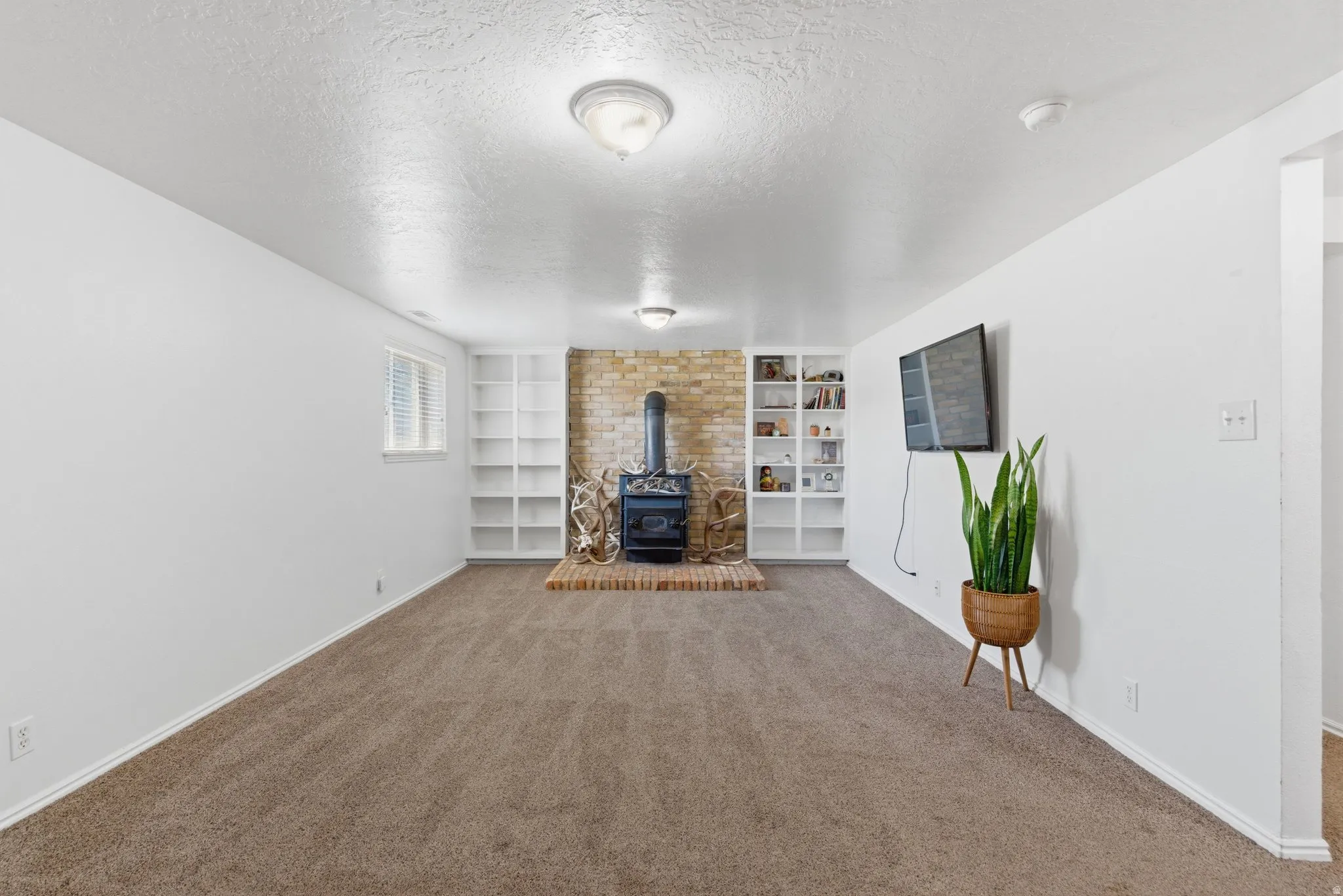 Spacious room featuring carpet flooring, built-in shelving, a brick accent wall, a wood-burning stove with brick hearth, and a window with blinds